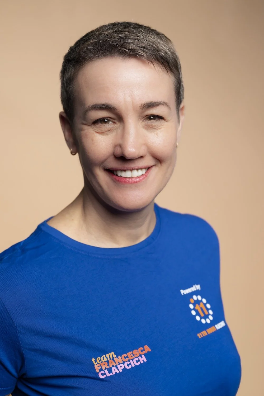 Smiling woman with short hair wearing a blue t-shirt with 'team FRANCESCA CLAPICH' printed on it, standing against a beige background.