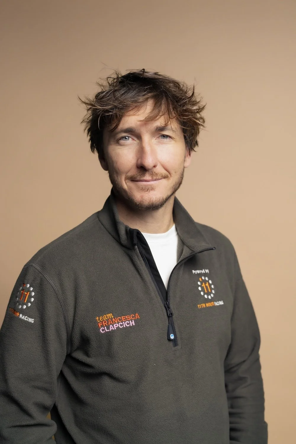 A man with brown hair, a short beard, and light skin, wearing a grey zip-up fleece with 'team Francesca Clapic' and racing logos, standing against a beige background, looking at the camera with a slight smile.