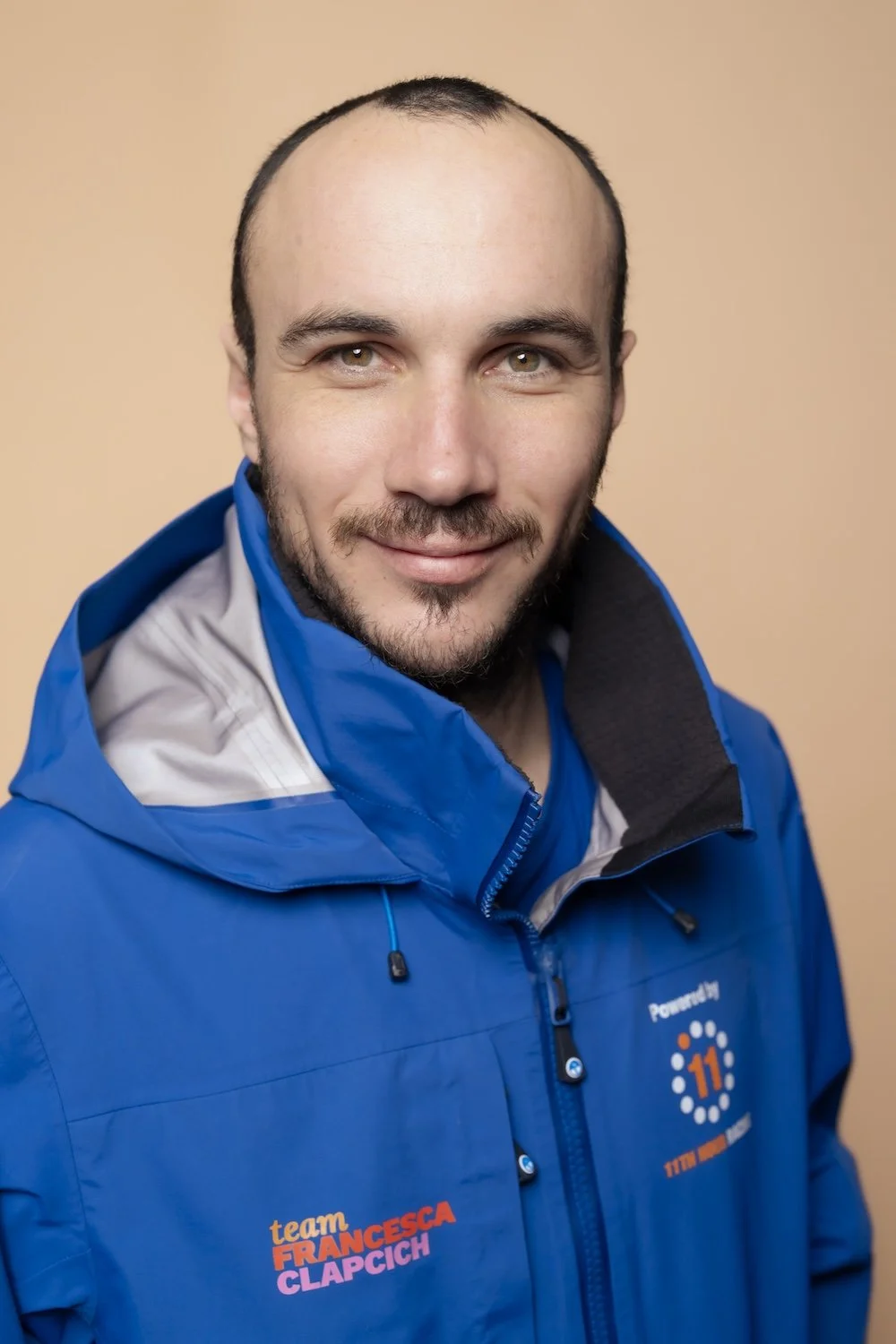A man with short hair and a beard, wearing a blue jacket with team logos, smiling at the camera against a beige background.