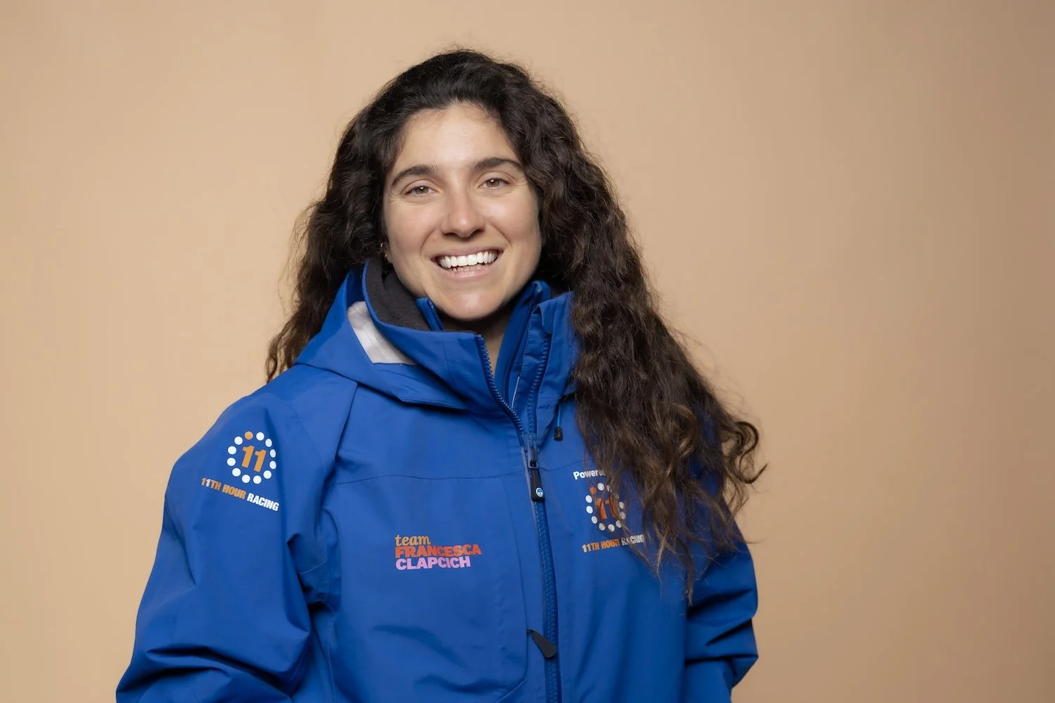 Smiling woman with long curly brown hair wearing a blue team jacket with logos, standing against a beige background.