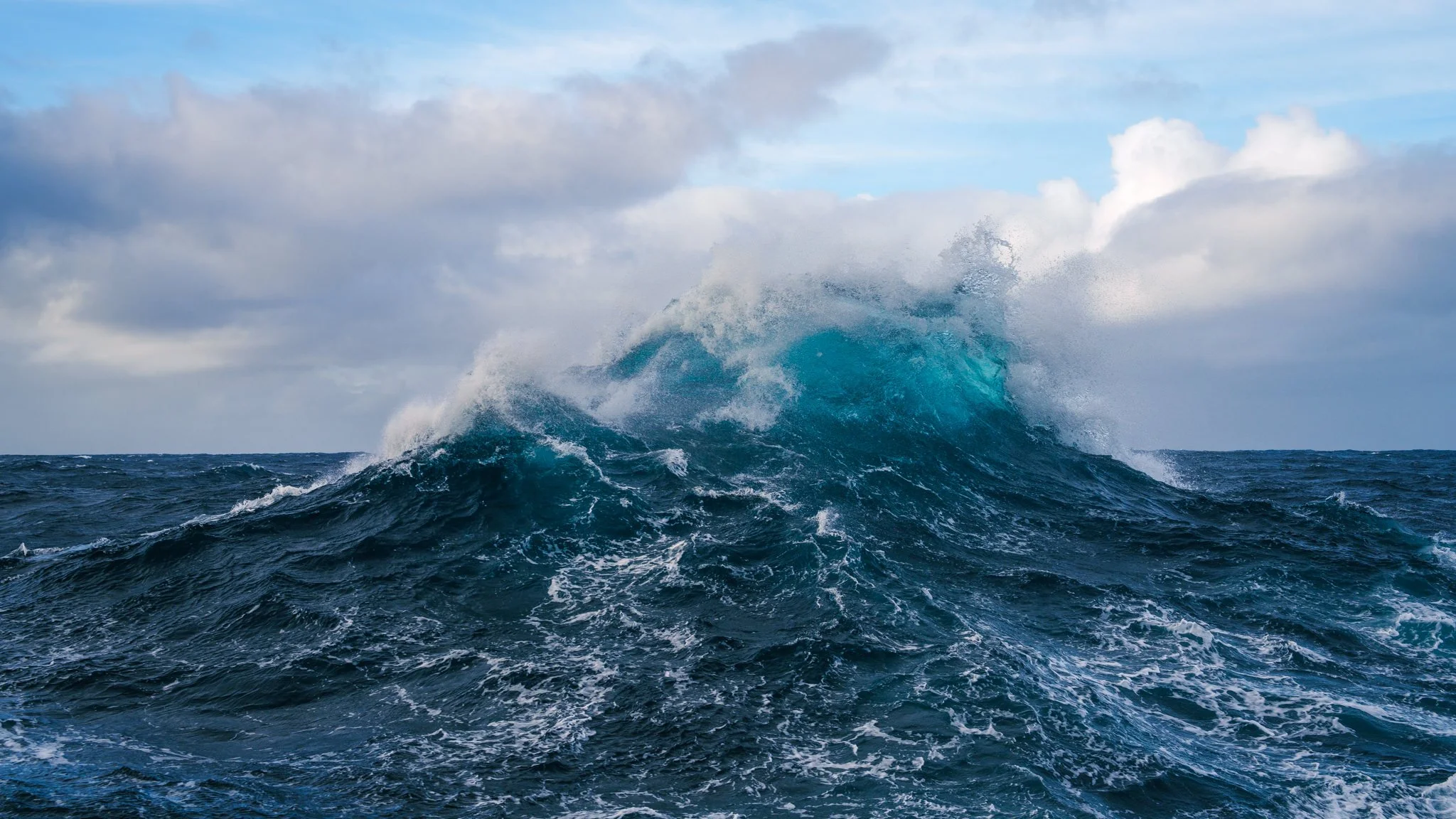 Le onde dell'oceano che si infrangono, con un'onda imponente in primo piano e un cielo nuvoloso sullo sfondo.