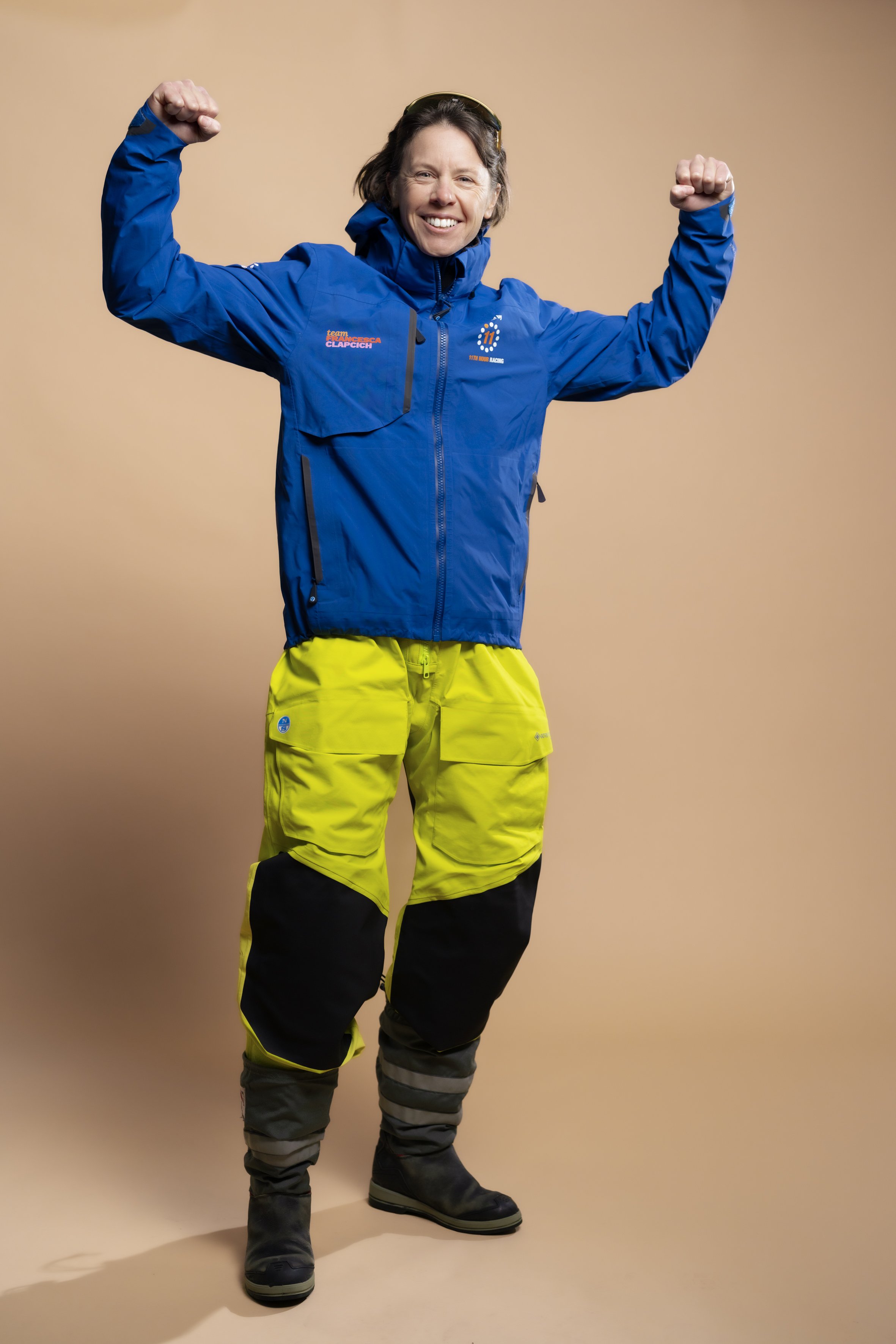 Person wearing a blue jacket, yellow athletic pants, and black boots, standing with arms raised in a victory pose against a beige background.