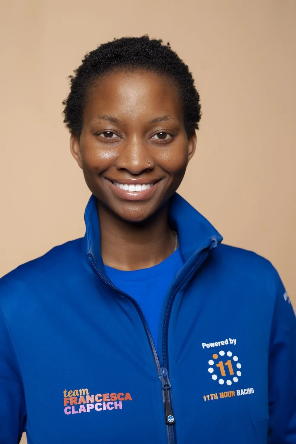 Portrait of a smiling woman with short curly hair wearing a blue jacket with logos and text, against a beige background.