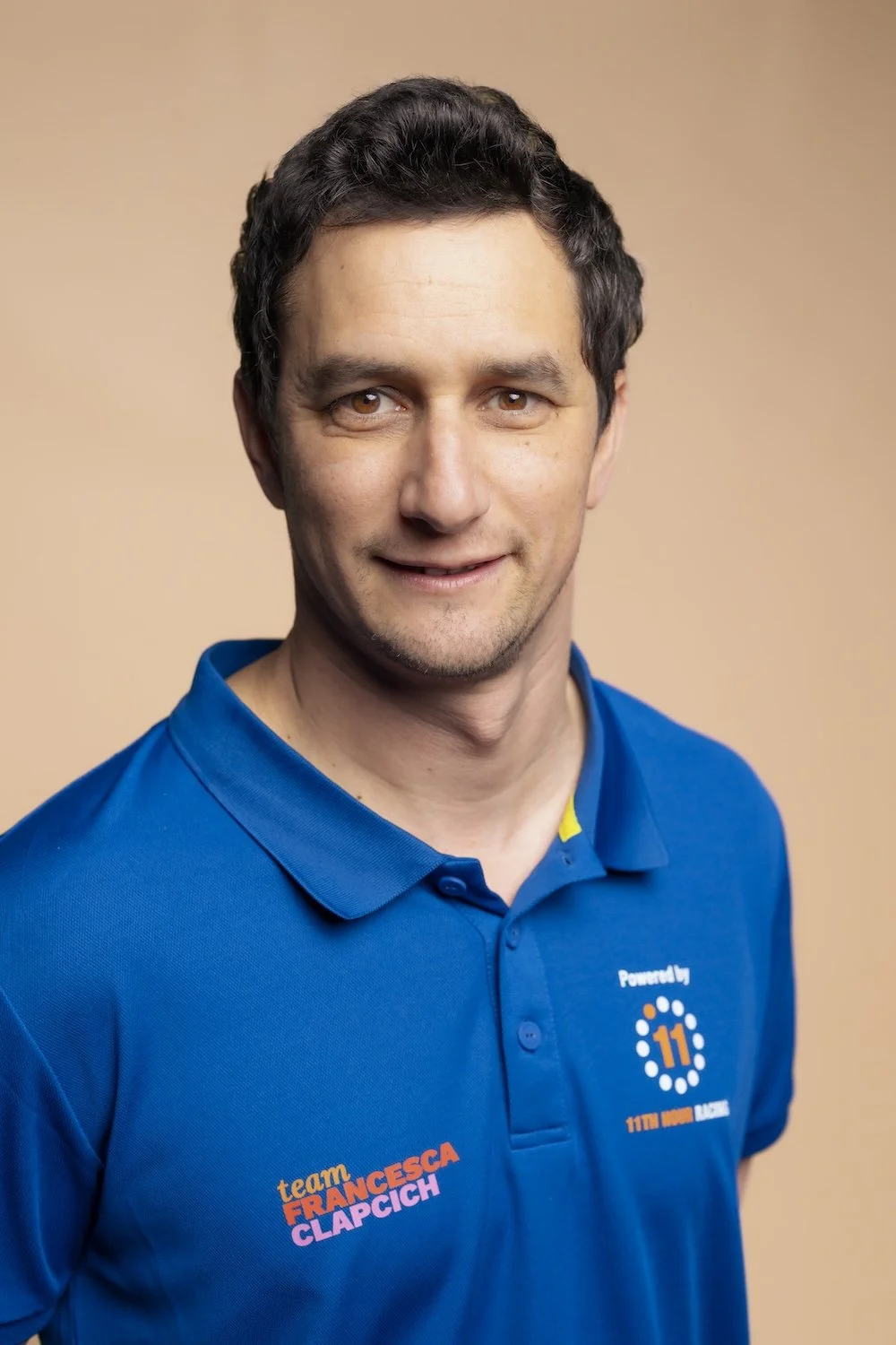Portrait of a man with short dark hair, wearing a blue polo shirt with team logos and text, smiling slightly, against a beige background.