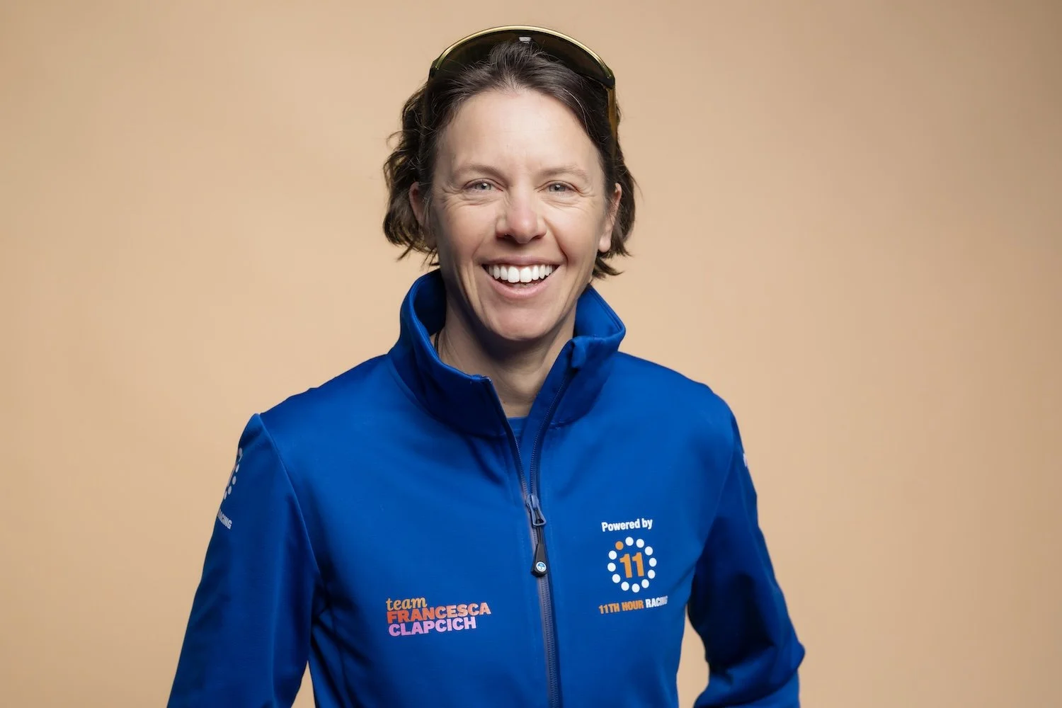 Smiling woman with short brown hair, wearing a blue zip-up jacket with logos for team Francesca Clapic and 11th Hour Racing, against a beige background.