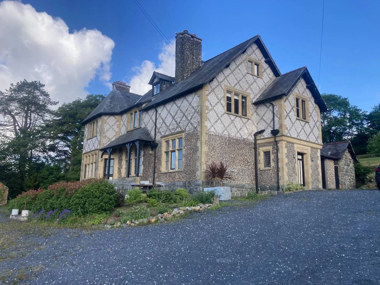 A large, historic stone house with a steep roof, multiple windows, and a chimney, surrounded by greenery and a gravel driveway under a partly cloudy sky.