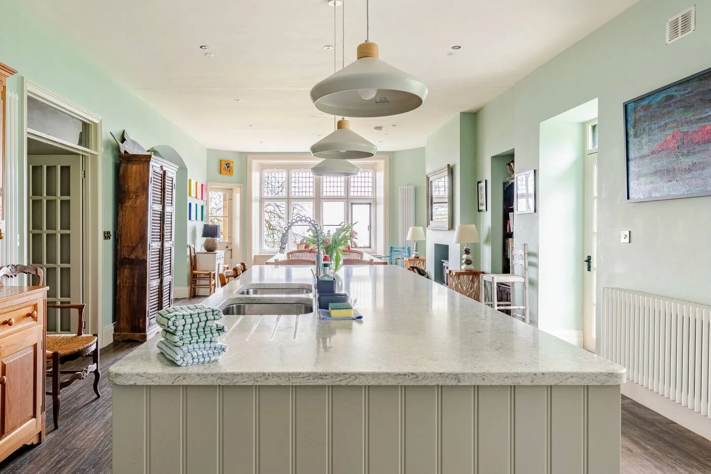 Bright kitchen with pale green walls, large central island with a white countertop, and pendant lights hanging above. There are various chairs and tables in the background, along with wall art and a windowsill with plants.