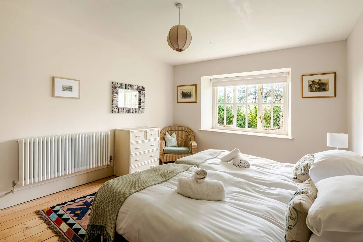 A bedroom with two unmade beds, white linens, rolled towels, a dresser, wicker chair, and a window showing greenery outside. Small framed pictures on the walls, a radiator, and a ceiling pendant light.