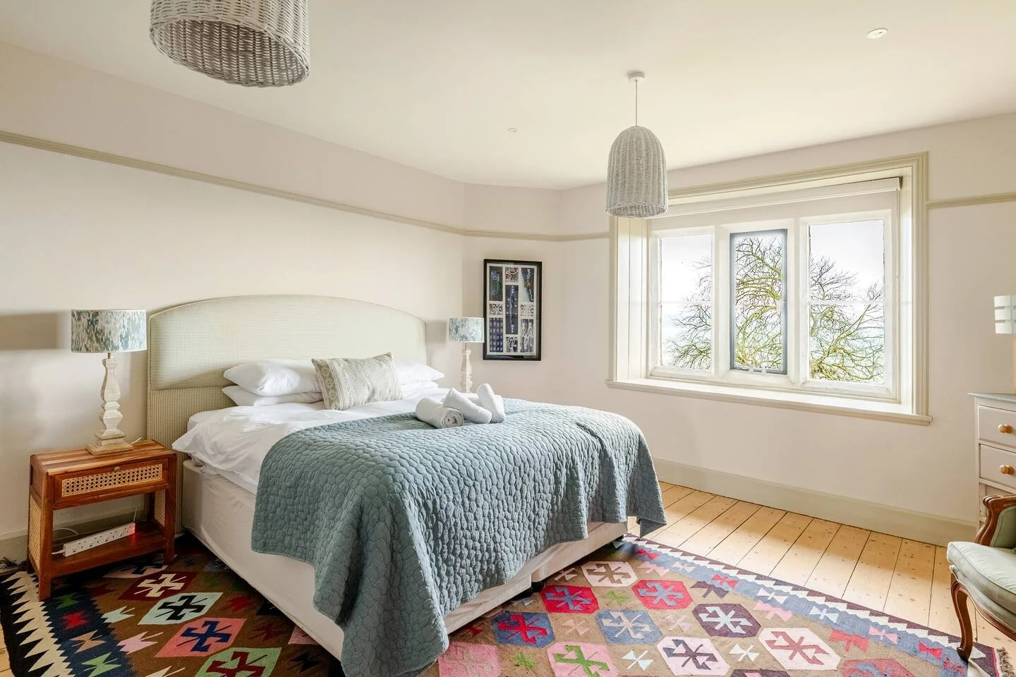 Bright bedroom with a large window, a bed with a light-colored headboard, blue quilt, and towels, two bedside lamps, colorful patterned rug, and a framed picture on the wall.