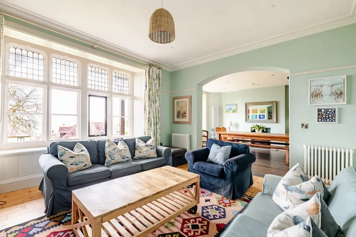 Living room with large window, three sofas with pillows, wooden coffee table, colorful rug, and art on walls.
