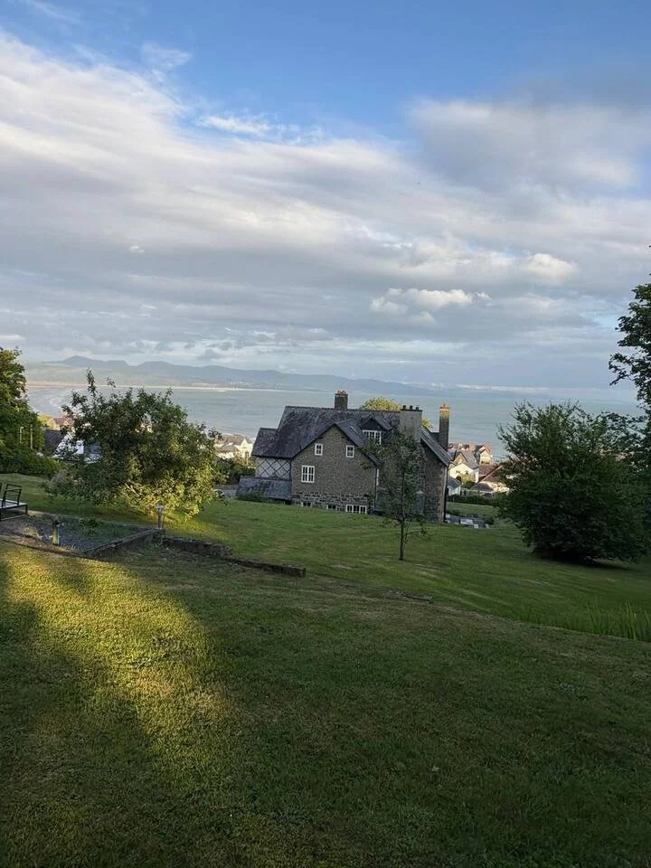 A scenic view of a gray stone house on a grassy hill with trees, overlooking a body of water under a partly cloudy sky.