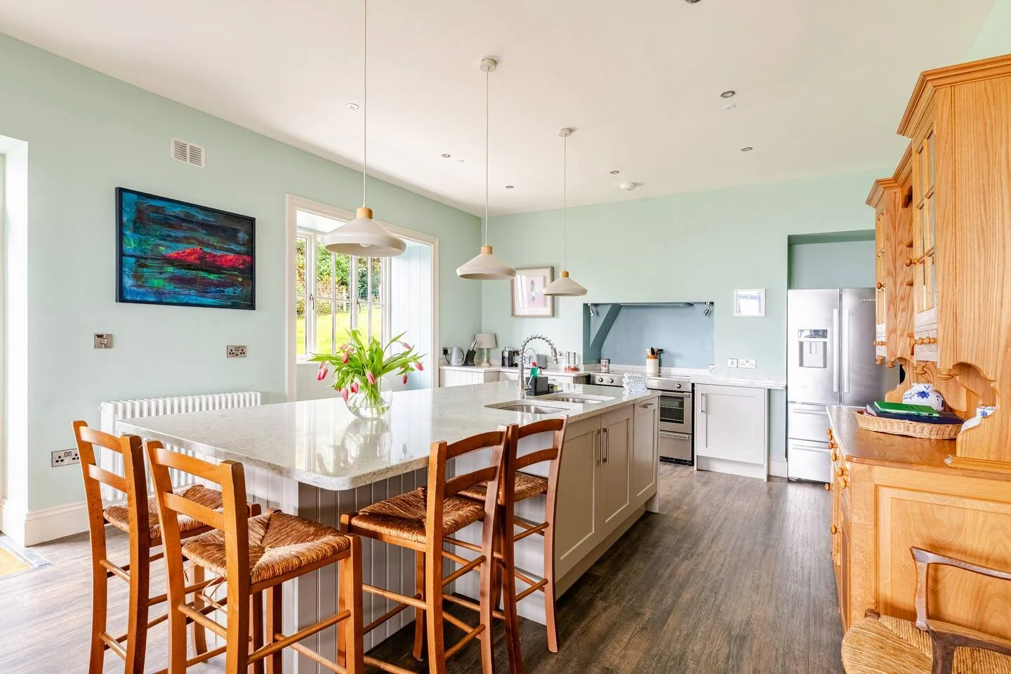 Bright kitchen with pastel green walls, wooden cabinetry, and a large island with a marble top. Three pendant lights hang above the island, which has a vase of flowers. The kitchen features modern appliances including a stainless steel refrigerator a