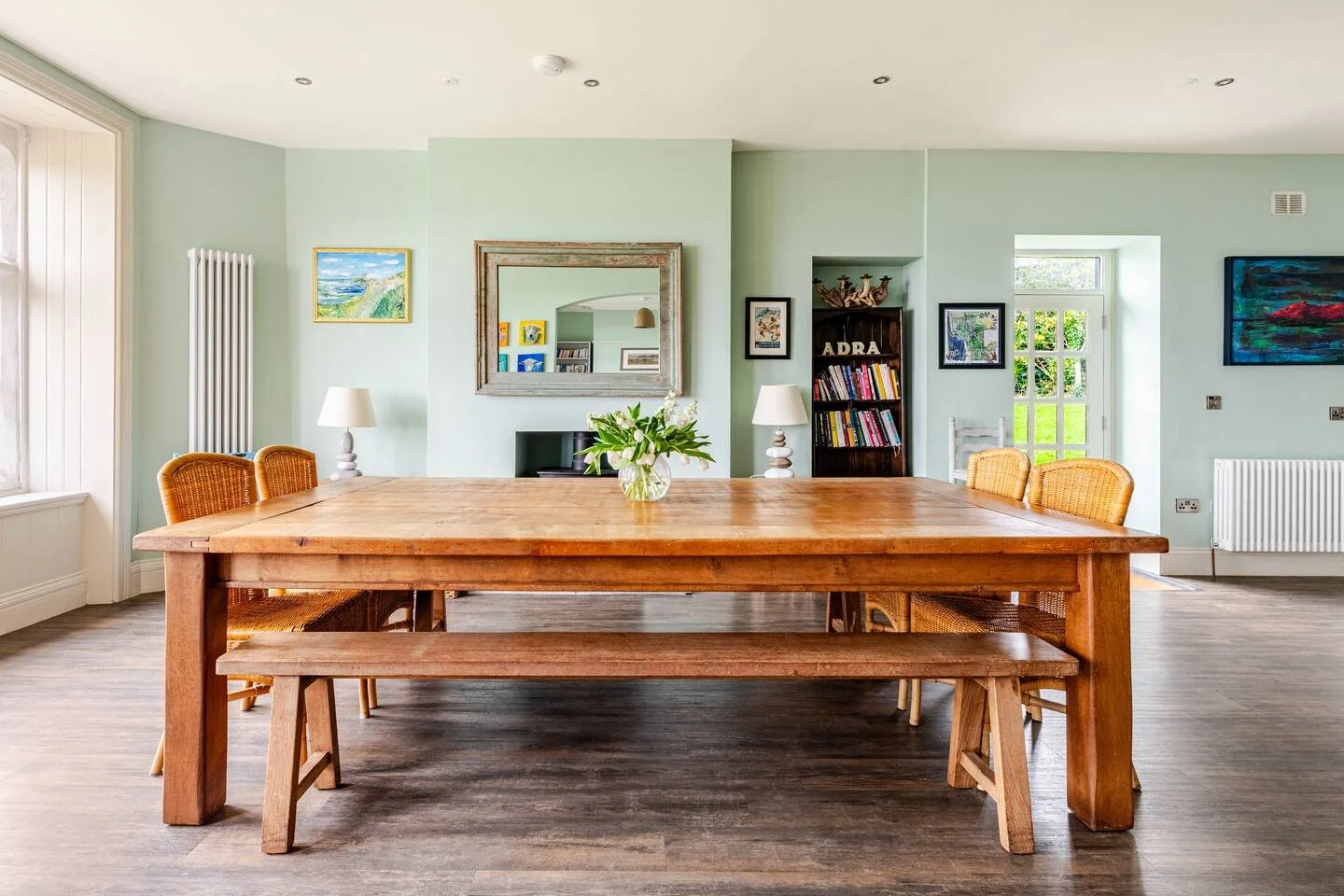 Bright dining room with wooden table, wicker chairs, paintings, mirror, lamps, bookshelf, and large windows with a garden view.