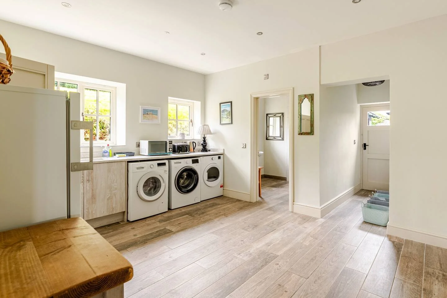 A bright laundry room with two windows, a countertop holding a microwave, toaster, and lamp, three laundry machines, and baskets near the door leading outside.