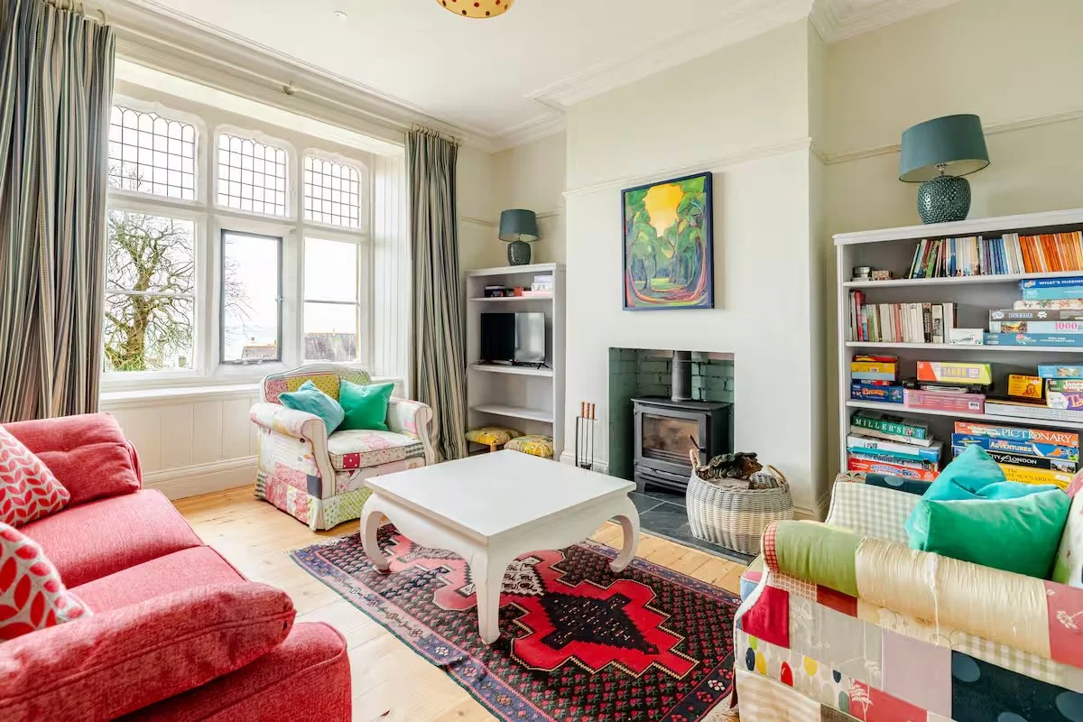 A cozy living room with colorful furniture, a fireplace, a bookshelf filled with books, and a window letting in natural light.