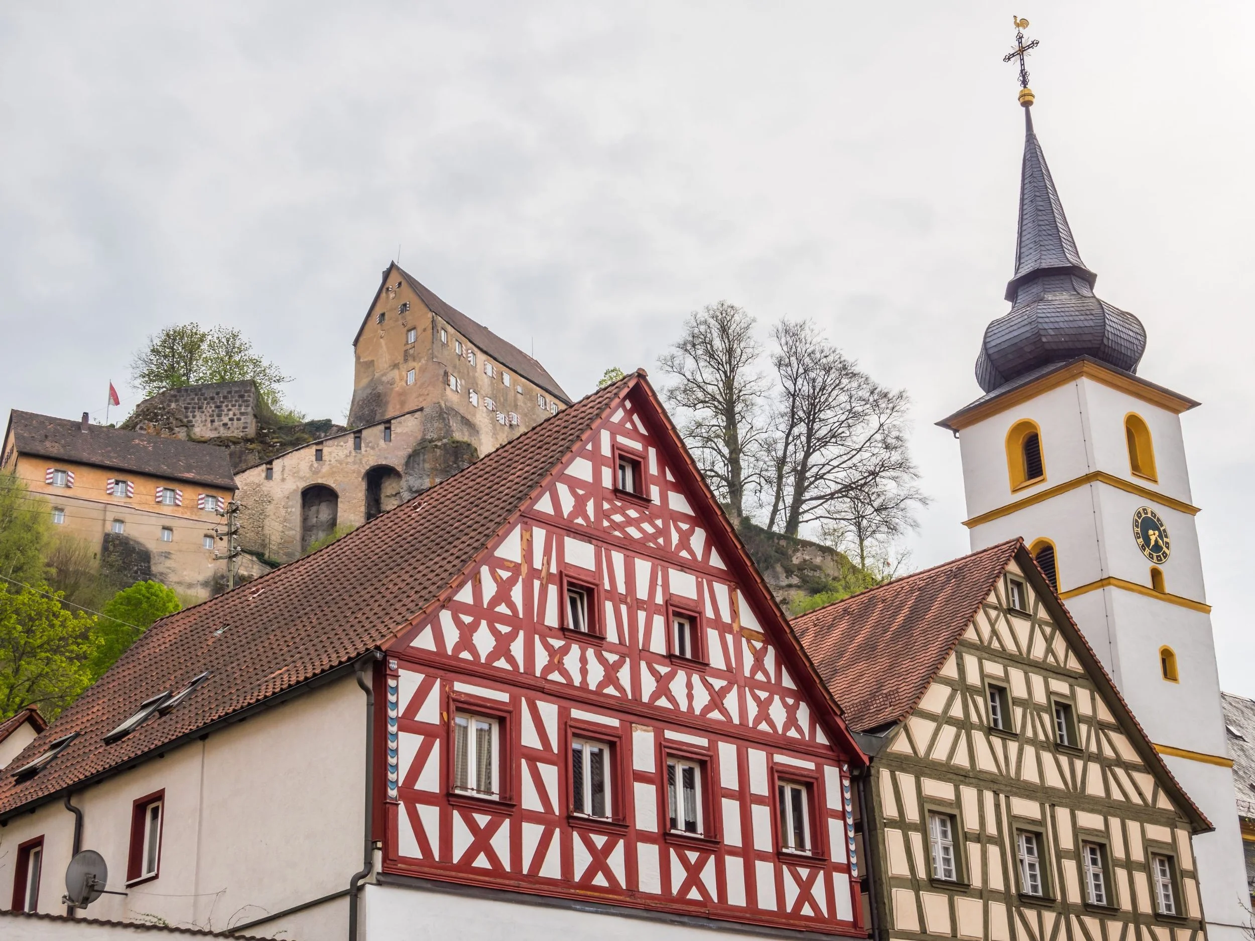 Historische Gebäude in einem malerischen europäischen Stadtviertel mit traditionellen Fachwerkhäusern, einer Kirche mit einem hohen Turm und einem Schloss auf einem Hügel im Hintergrund, umgeben von Bäumen.