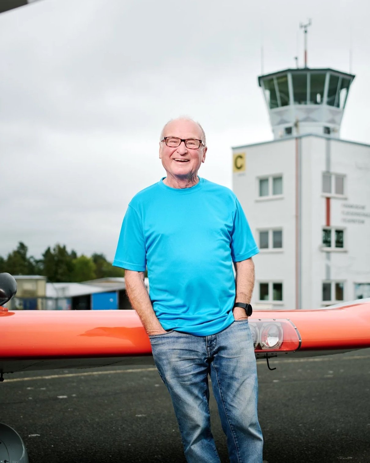 Ein älterer Mann in blauer T-Shirt und Jeans steht vor einem kleinen Flugzeug auf einem Flugfeld, im Hintergrund ein Kontrollturm.