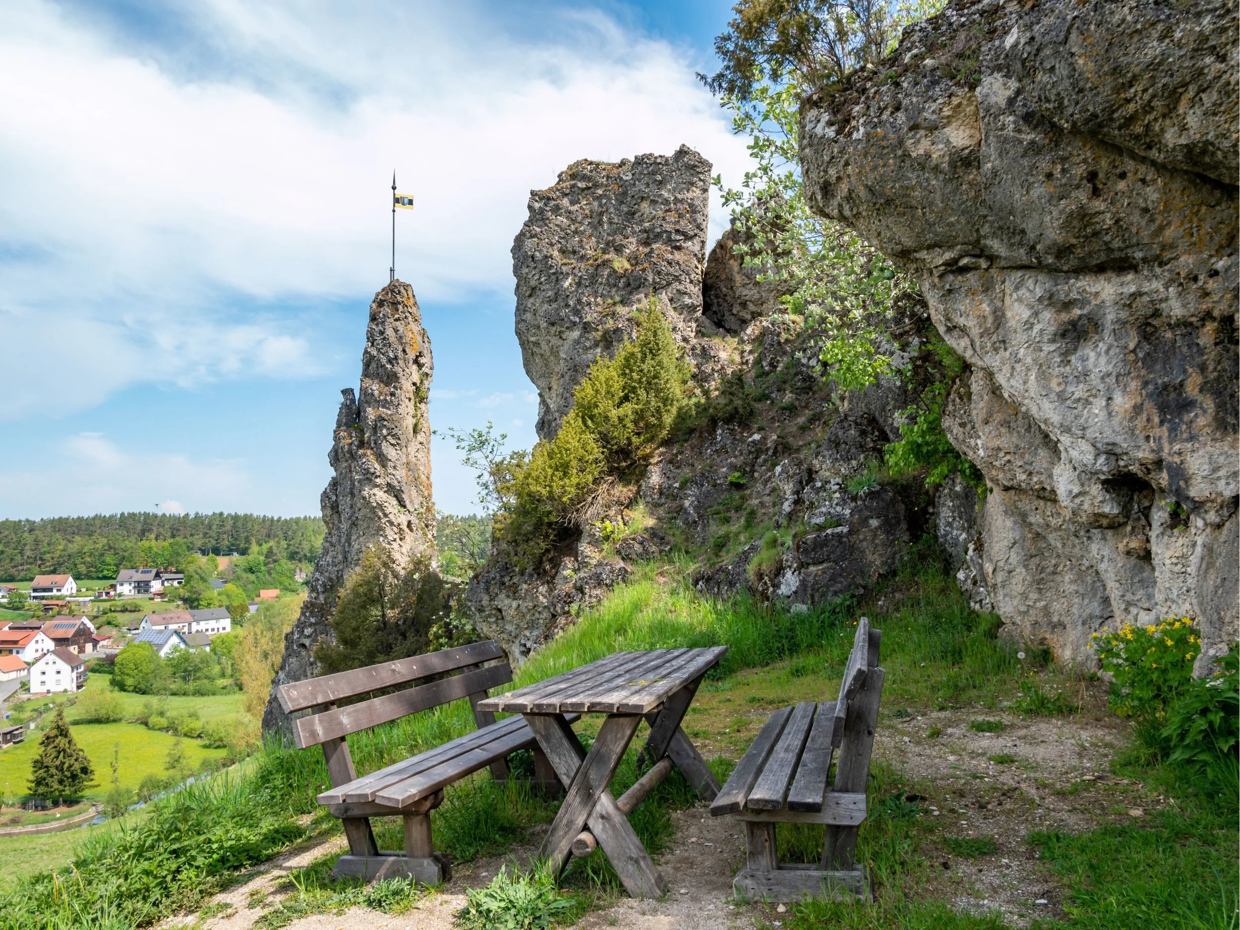 Zwei Holzsitzecke vor einer großen Felswand mit Büschen und Bäumen, Blick auf eine grüne Landschaft mit Häusern und Wäldern, Himmel mit Wolken.