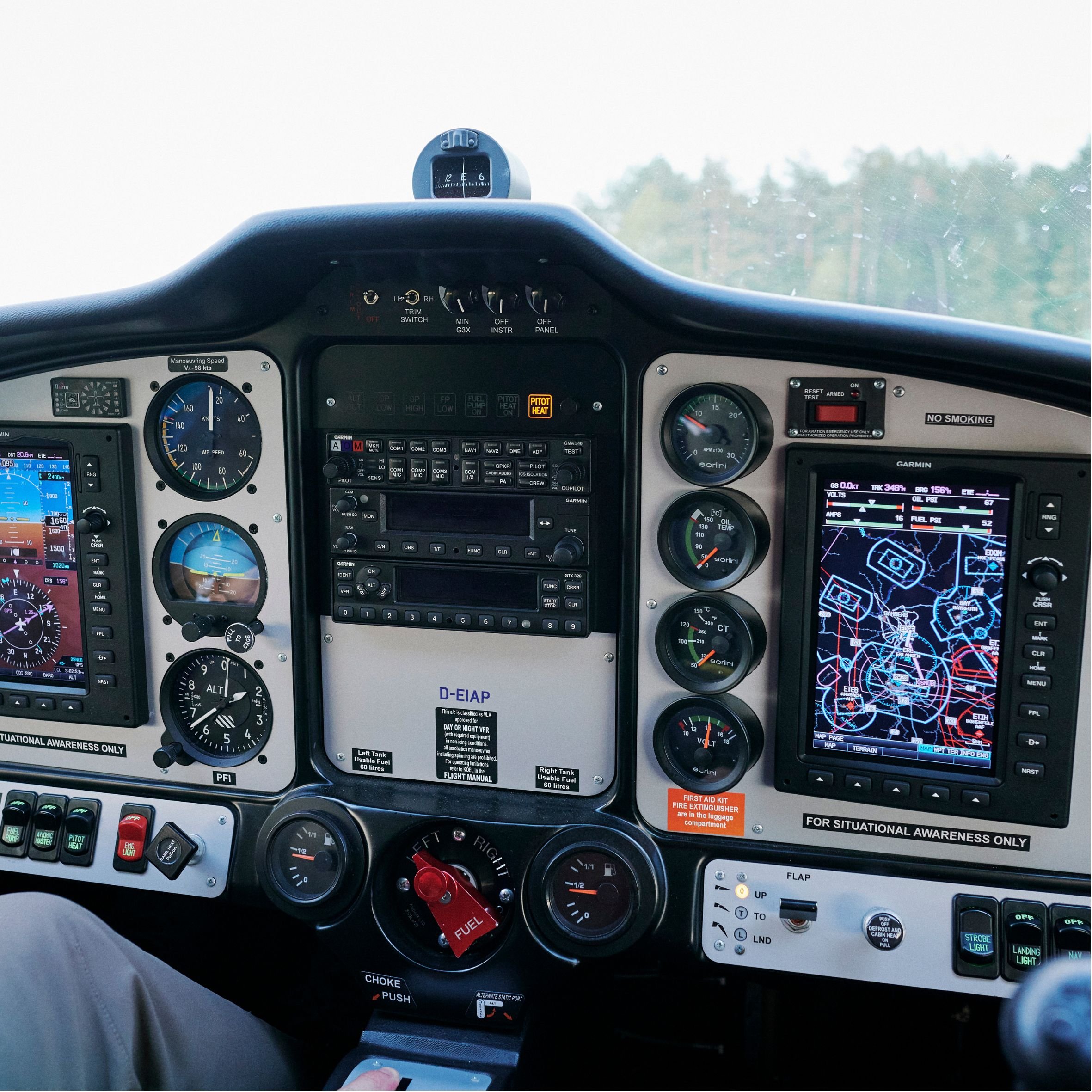 Innensicht eines Flugzeugcockpits mit verschiedenen Anzeigen, Steuerknüppeln, Instrumenten und Bildschirmen, Blick nach vorne auf das Flugfeld und das Wetter außerhalb.
