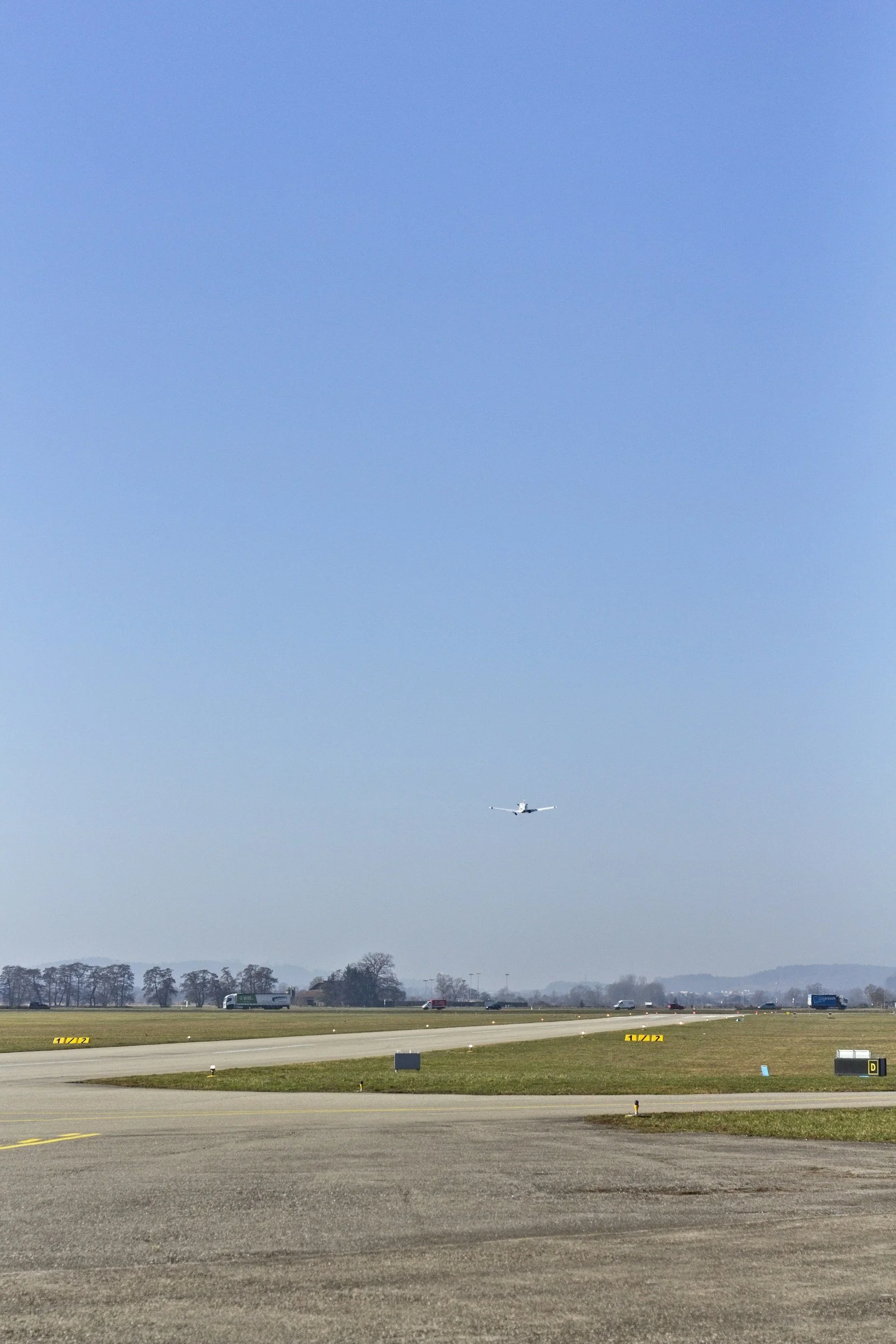 Ein Flugzeug fliegt hoch am Himmel über einer Start- und Landebahn eines Flughafens mit Bussen und Panoramabus auf dem Boden. Im Hintergrund sind Bäume und eine klare Himmel zu sehen.