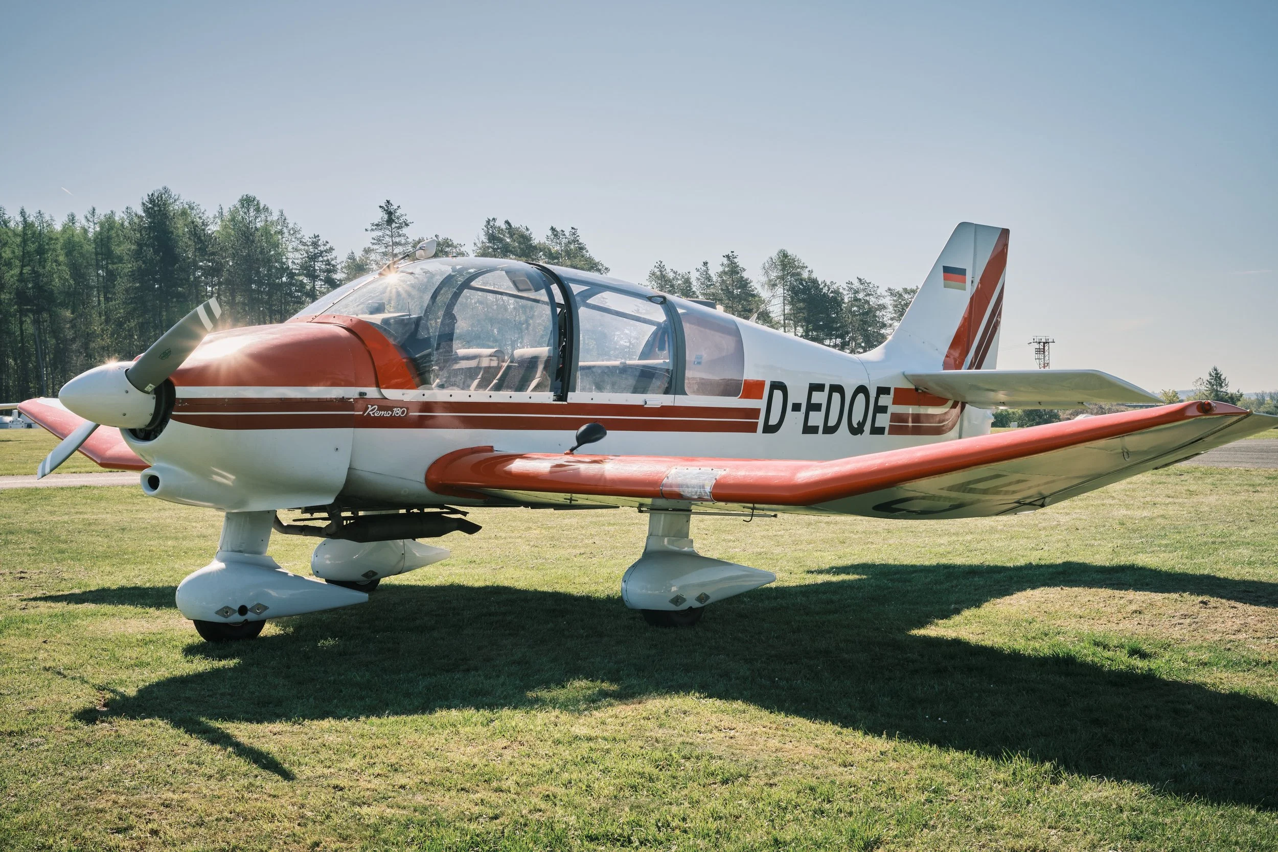 Ein weiß-rotes Sportflugzeug auf einer Wiese, im Hintergrund Bäume und ein blauer Himmel.