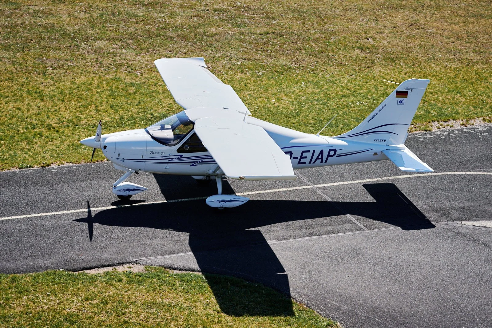 Ein weißes Kleinflugzeug steht auf einem Flugplatz neben einer Grasfläche, mit seinem Schatten auf dem Asphalt.