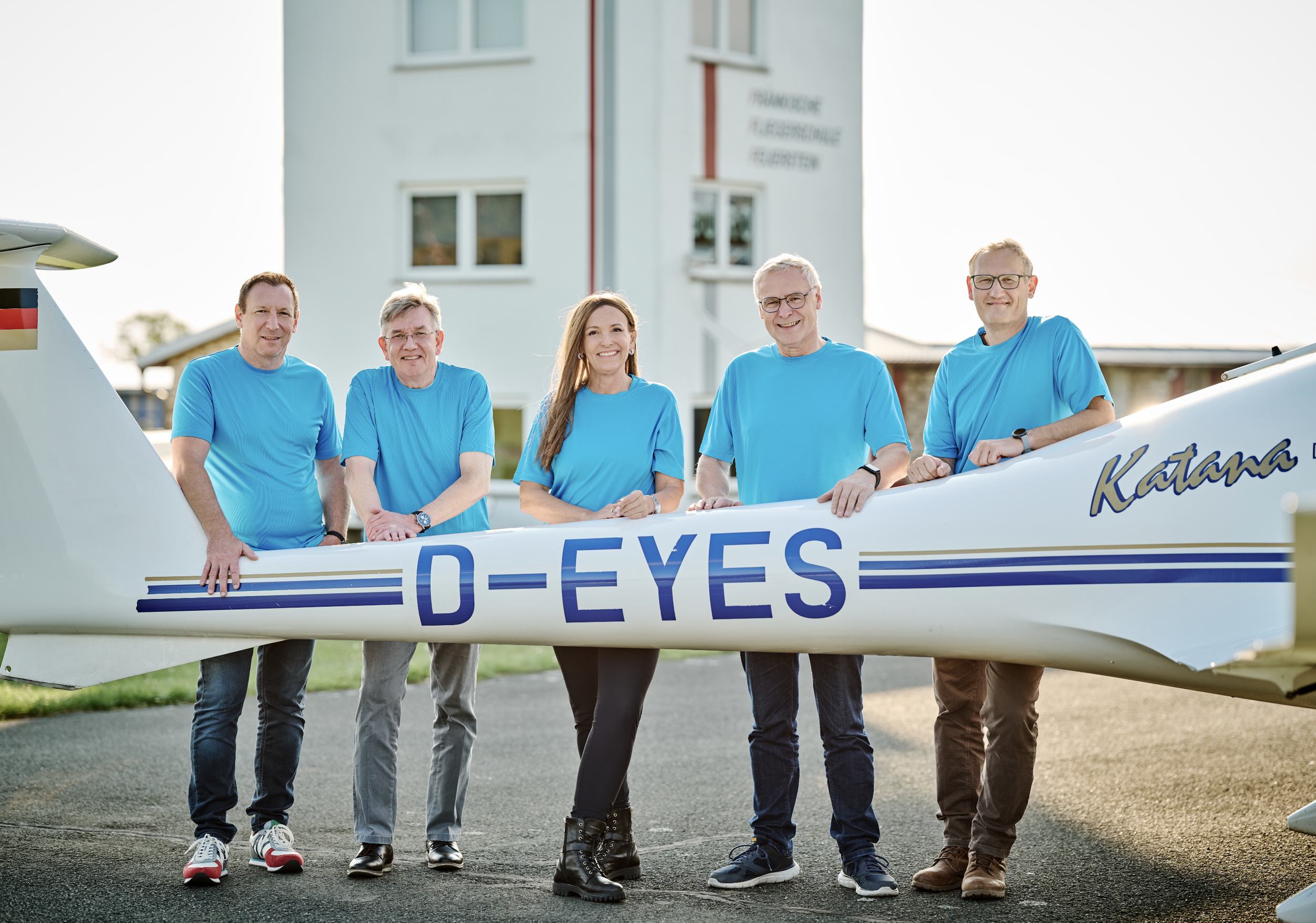 Fünf Personen in blauen Shirts stehen vor einem Flugzeug mit der Aufschrift »D-EYES«. Im Hintergrund ein Mehrfamilienhaus.