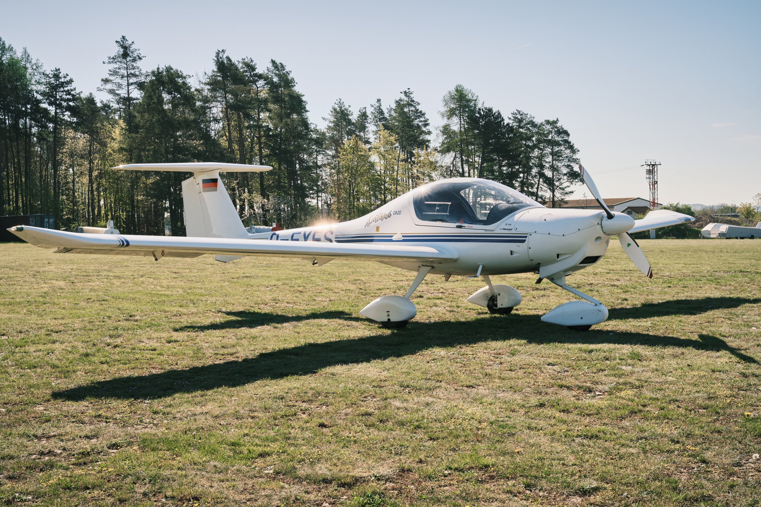 Ein kleines weißes Flugzeug steht auf einer Grasfläche, im Hintergrund Bäume und Häuser, bei sonnigem Wetter.