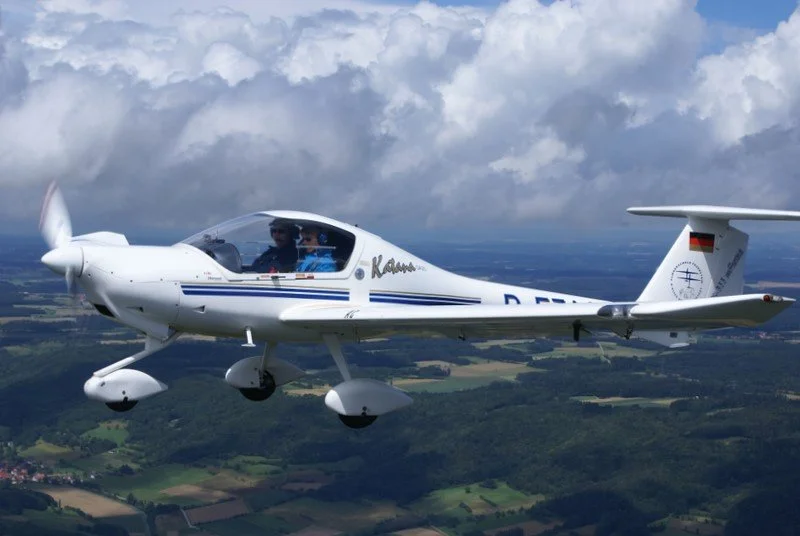 Ein weißes Privatflugzeug in der Luft mit Piloten im Cockpit über einer grünen Landschaft mit Wolken im Himmel.