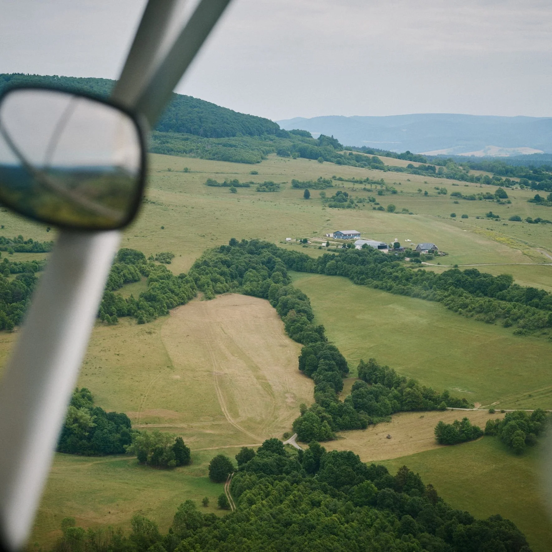 Landschaft mit grünen Hügeln, Bäumen, einigen Häusern und farmen, aufgenommen aus einem Flugzeug, Fenster im Vordergrund.