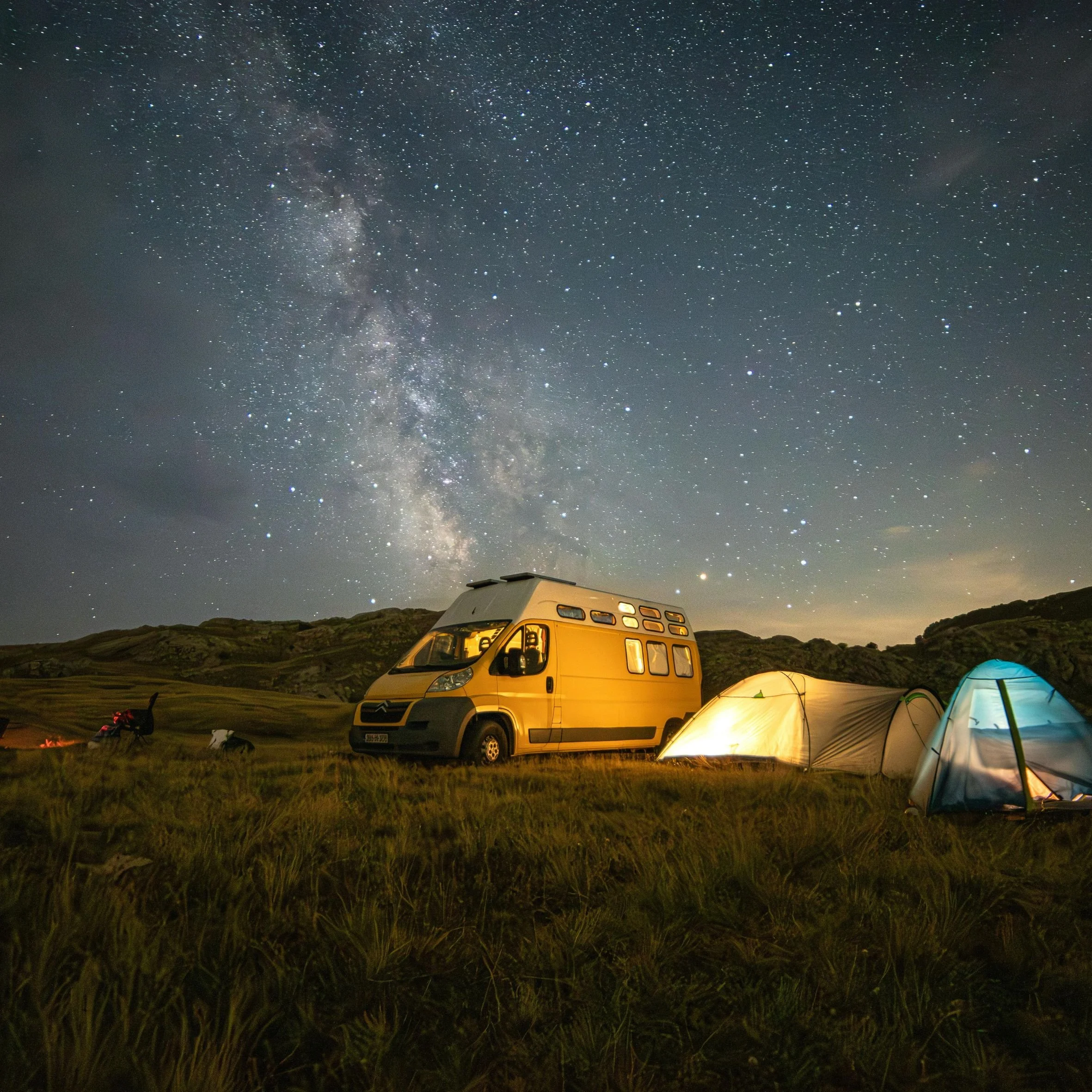Campingplatz bei Nacht mit Zelten, einem Wohnmobil und Sternenhimmel, darunter eine grüne Wiese in bergigem Gelände.