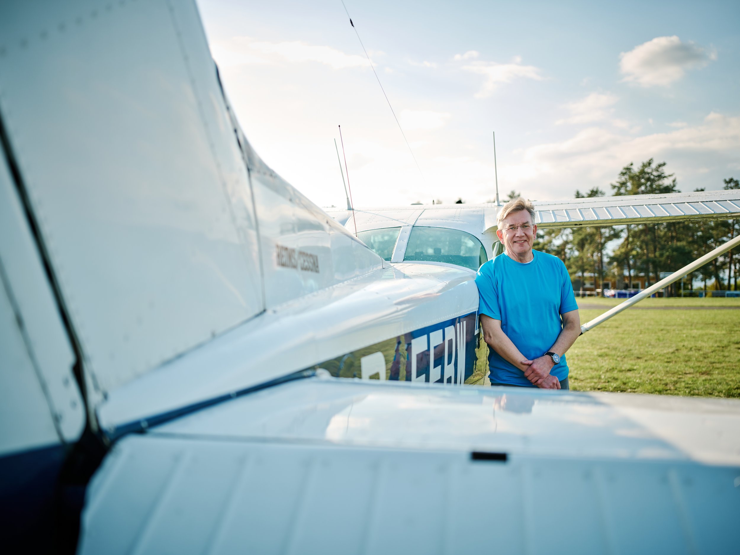 Ein Mann in blauer T-Shirt steht vor einem kleinen weißen Flugzeug im Freien, im Hintergrund sind Bäume und ein blauer Himmel mit Wolken.