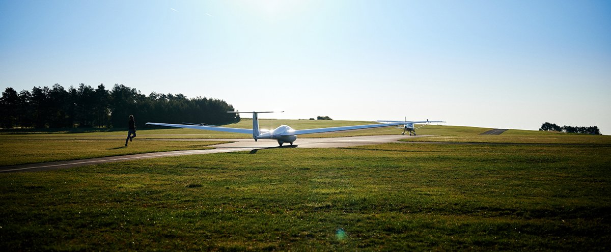 Ein Flugzeug steht auf einer startbereiten Landebahn, im Hintergrund sind Bäume und eine Person läuft vorbei, bei sonnigem Himmel.