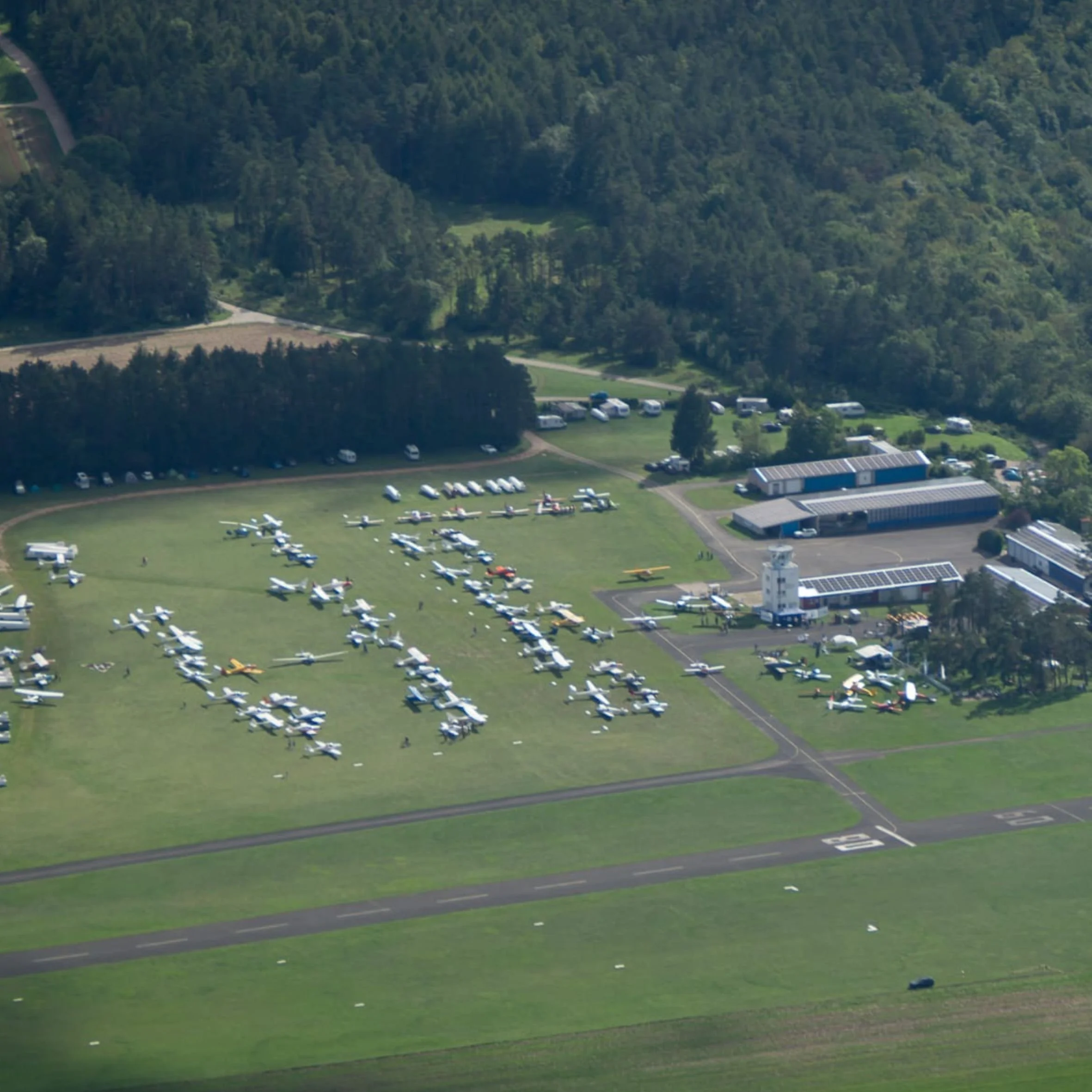 Luftaufnahme eines Flugplatzes mit vielen kleinen Flugzeugen, umgeben von Wäldern und einigen Gebäuden.