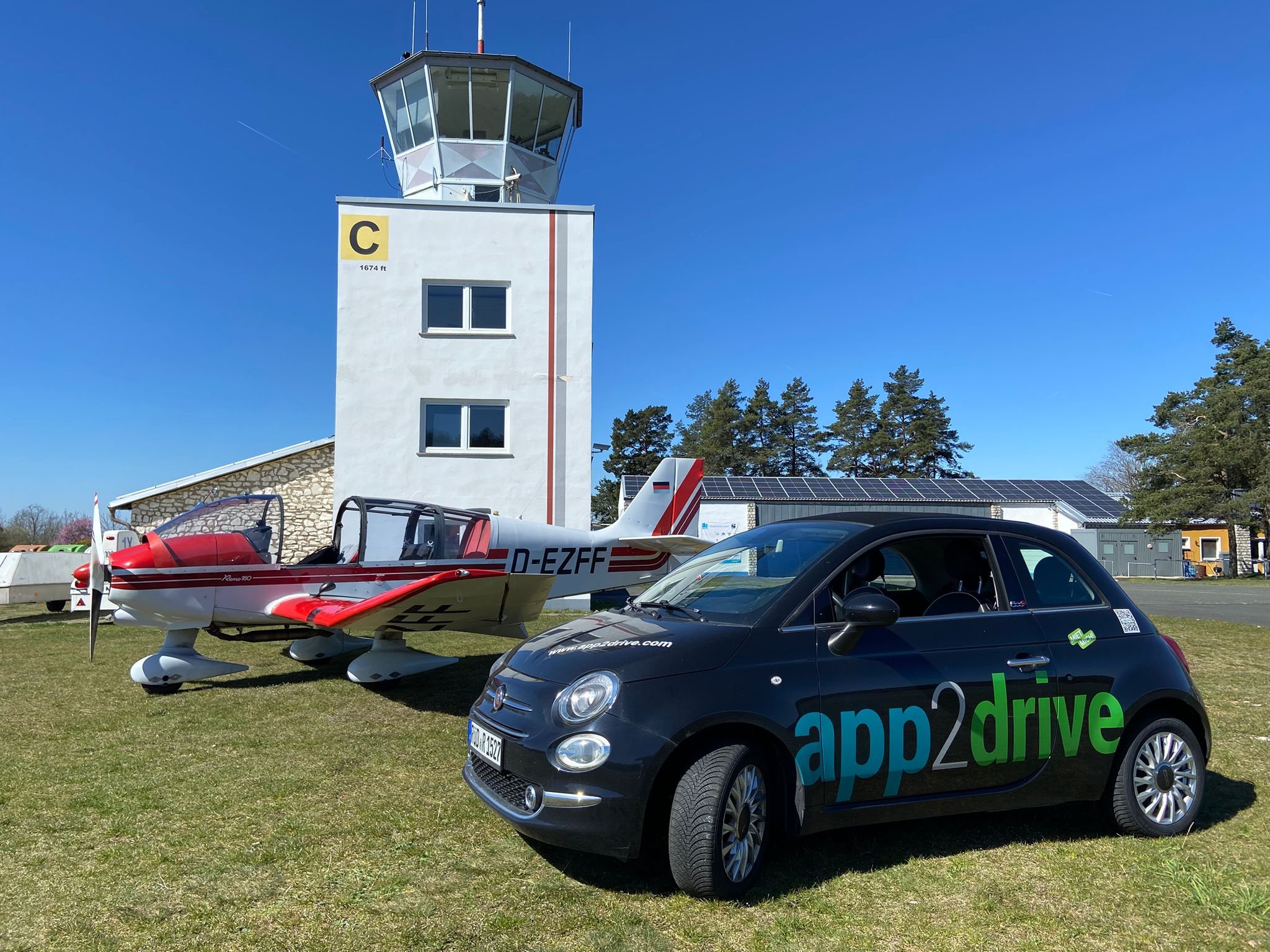 Ein Flugzeug und ein Auto stehen auf einer Wiese vor einem Kontrollturm mit blauem Himmel im Hintergrund.