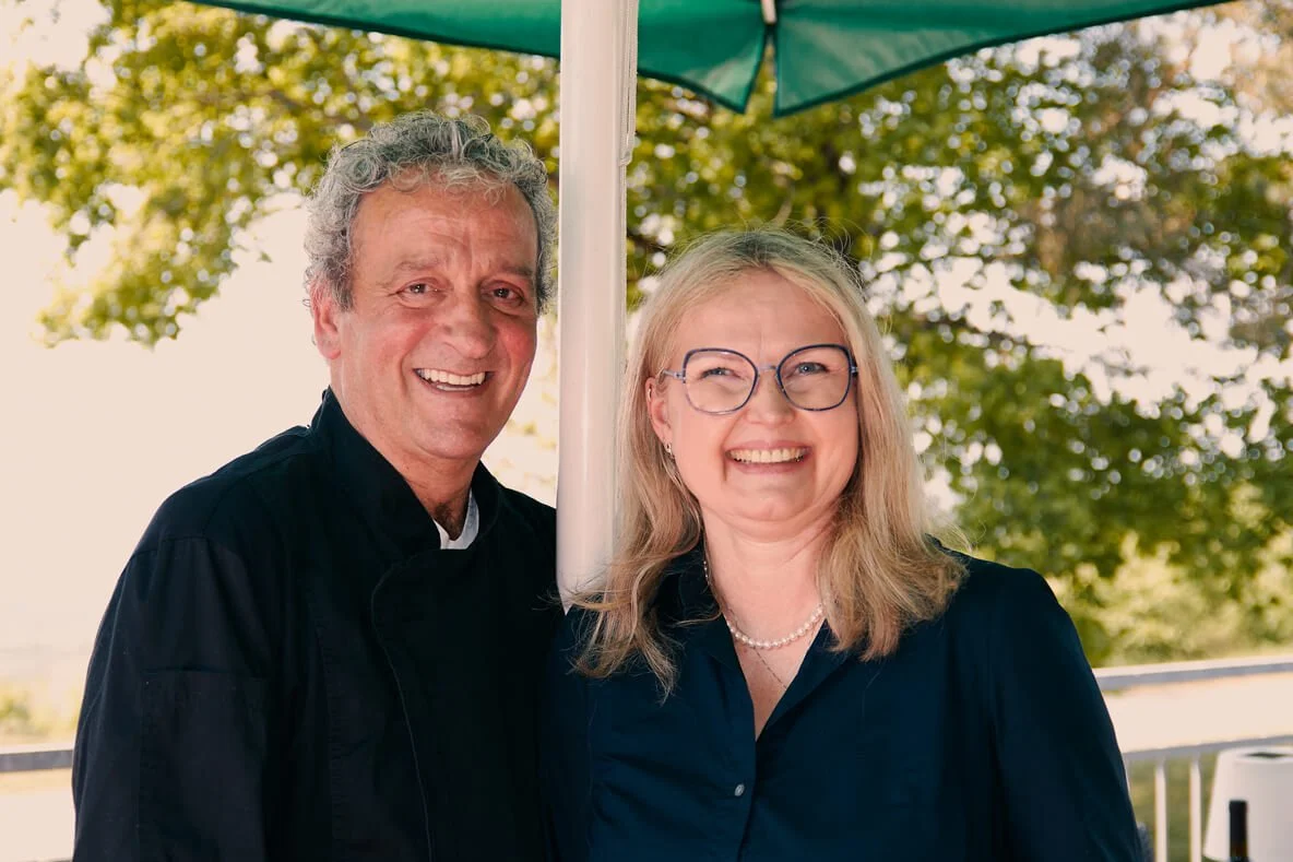 Ein Mann und eine Frau stehen unter einem grünen Sonnenschirm im Freien und lachen. Im Hintergrund sind Bäume mit grünen Blättern.