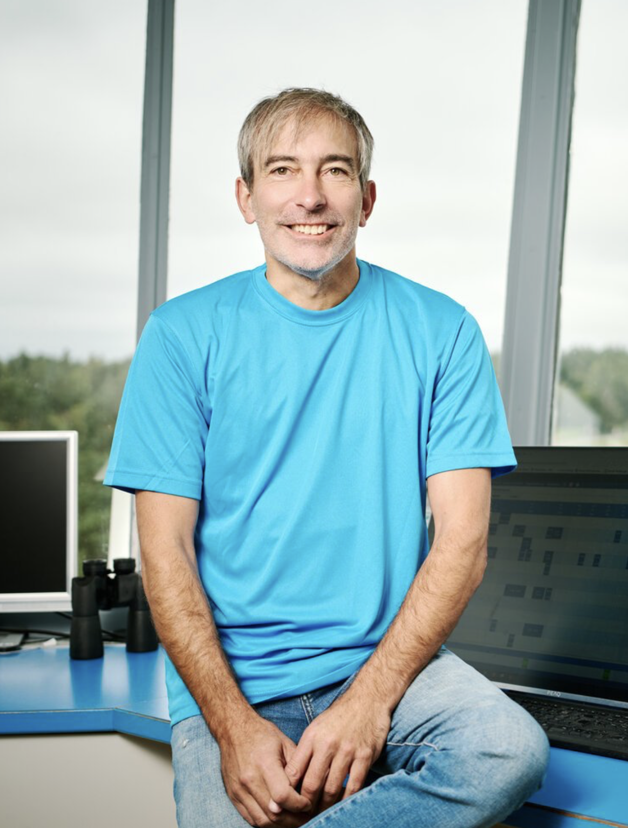 Ein Mann mit grauem Haar und Bart sitzt in einem modernen Büro, trägt ein hellblaues T-Shirt und lächelt, im Hintergrund große Fenster und Computerbildschirme.