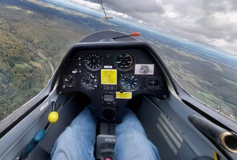 Innenaufnahme eines Segelflugzeugcockpits mit Blick nach vorne auf die Instrumententafel, while der Pilot sitzt. Draußen sind Wolken und Landschaft sichtbar.