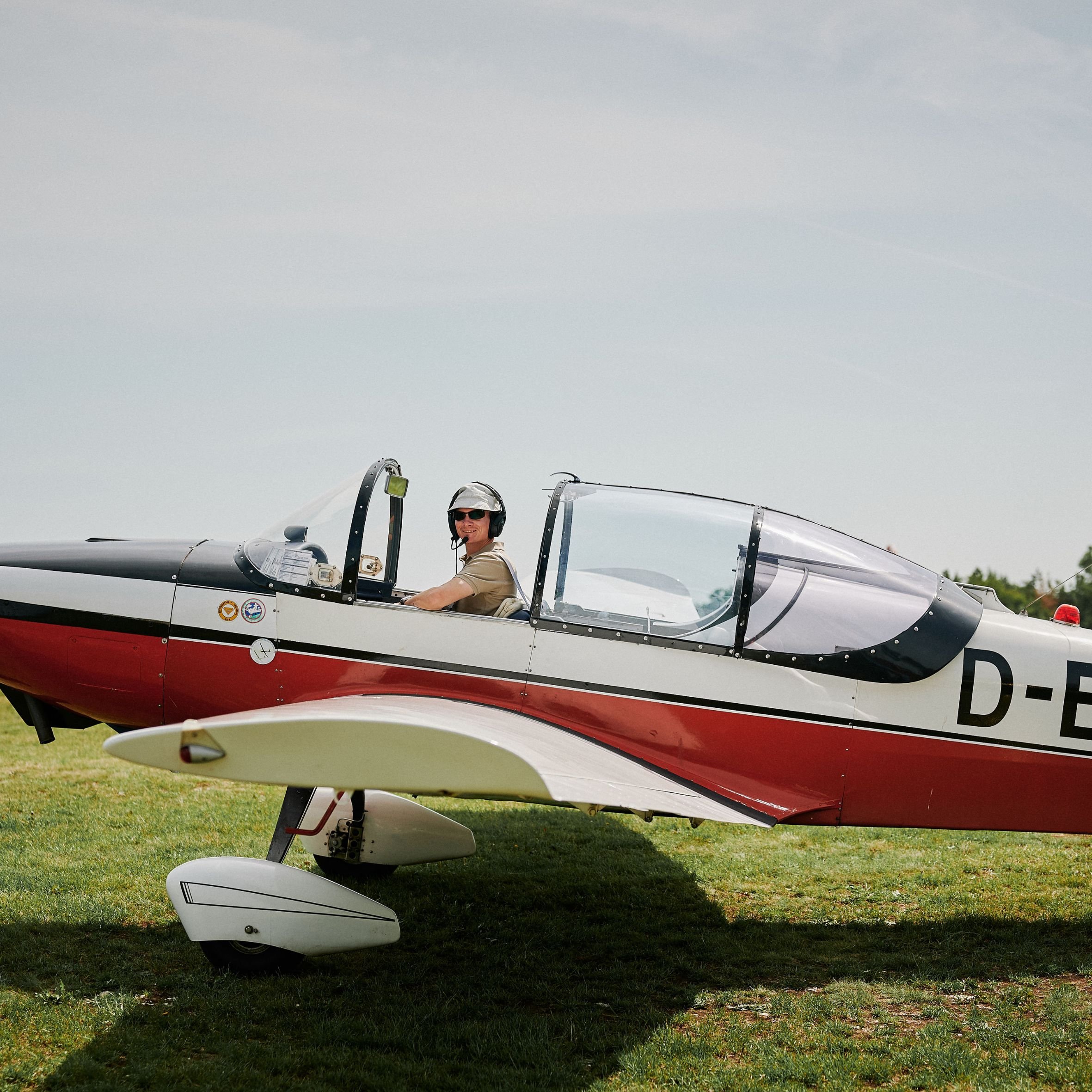 Ein Mann sitzt im Cockpit eines Kampfflugzeugs, trägt einen Helm und eine Sonnenbrille, im Hintergrund grauer Himmel.