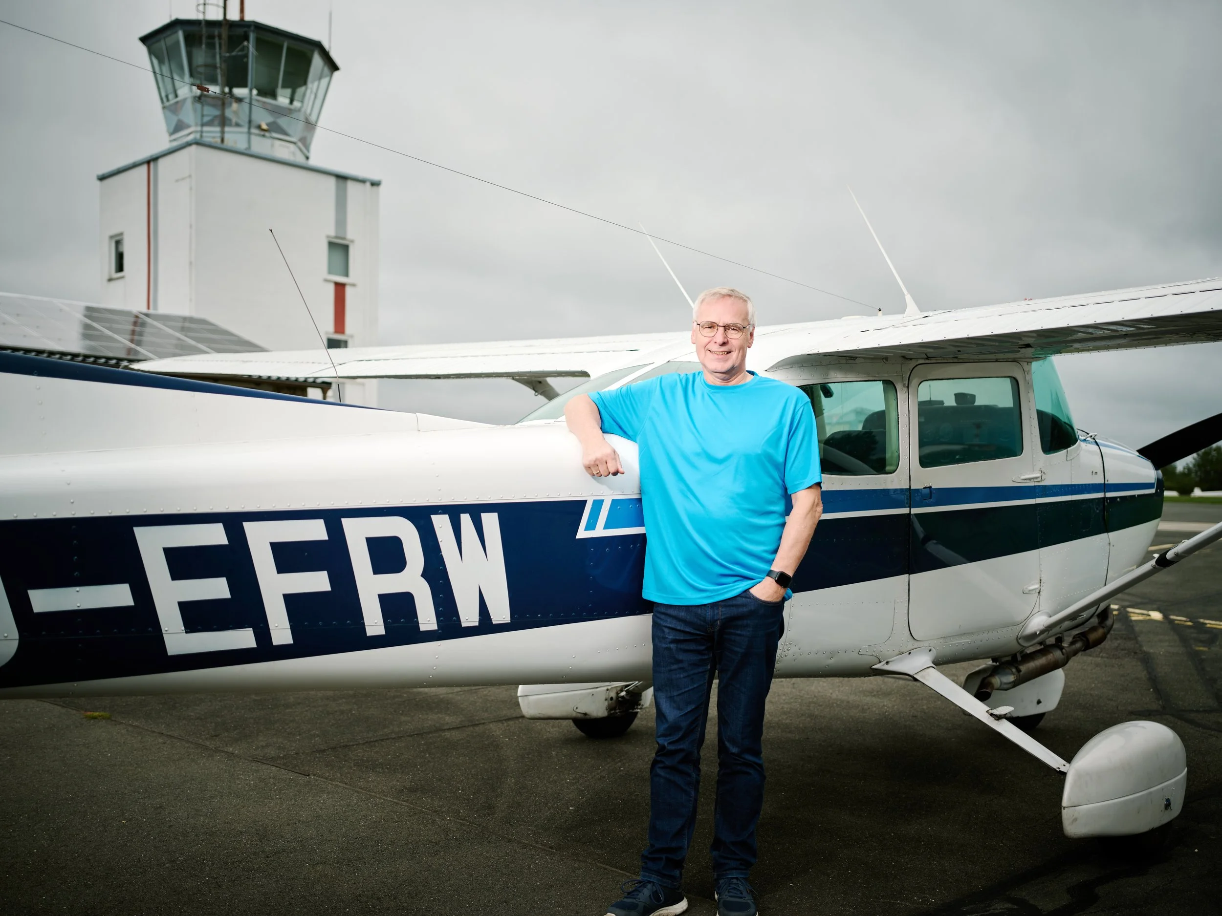Ein älterer Mann in einem blauen T-Shirt steht vor einem kleinen weiß-blauen Flugzeug auf einem Flugplatz. Im Hintergrund ist eine Kontrollturm zu sehen. Das Wetter ist bewölkt.