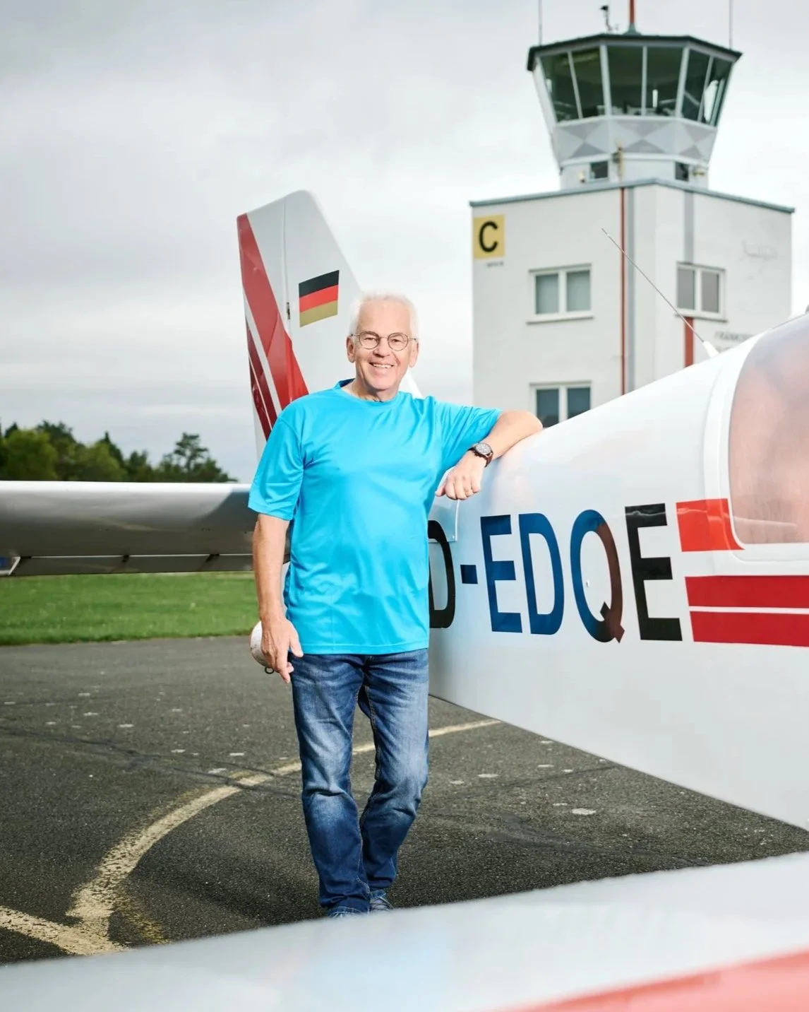 Ein älterer Mann in blauer T-Shirt steht neben einem Flugzeug auf einem Flughafen, im Hintergrund ein Kontrollturm.