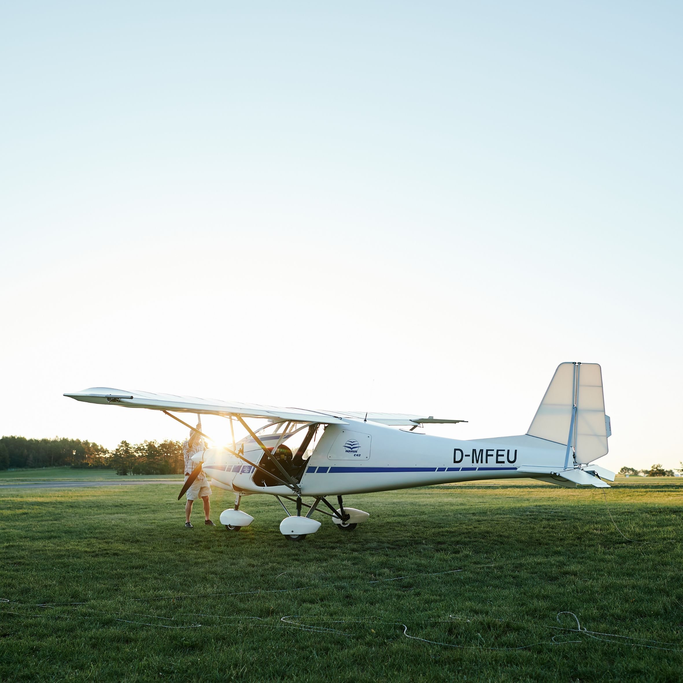 Ein kleines Flugzeug mit deutschem Kennzeichen parkt auf einem Rasen, während Menschen in der Nähe den Propeller vorbereiten, bei Sonnenuntergang.