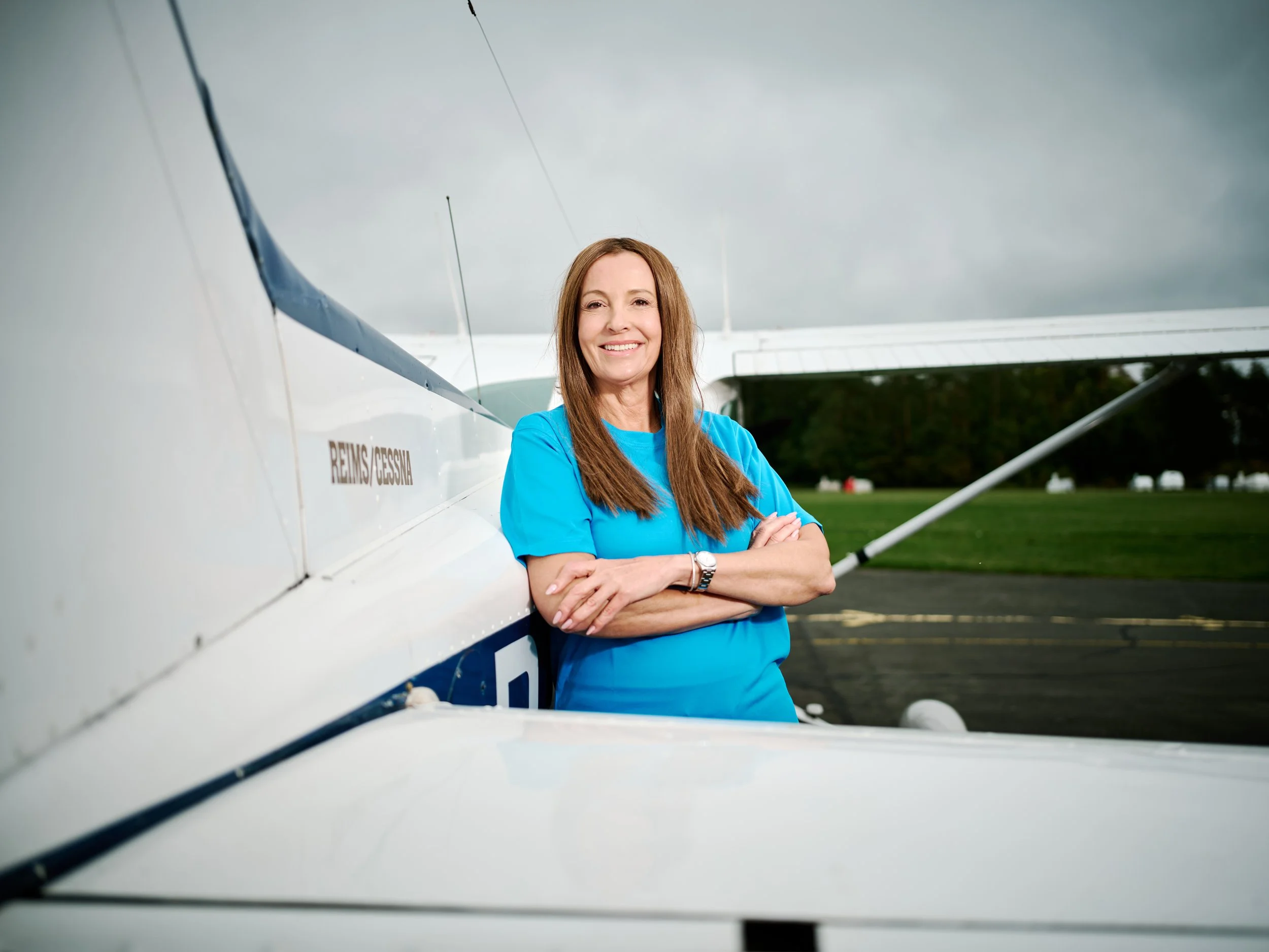 Eine lachende Frau mit braunen Haaren in blauer Sportbekleidung steht vor einem kleinen Flugzeug auf einem Flugfeld, im Hintergrund sind Bäume und Wolken am Himmel sichtbar.