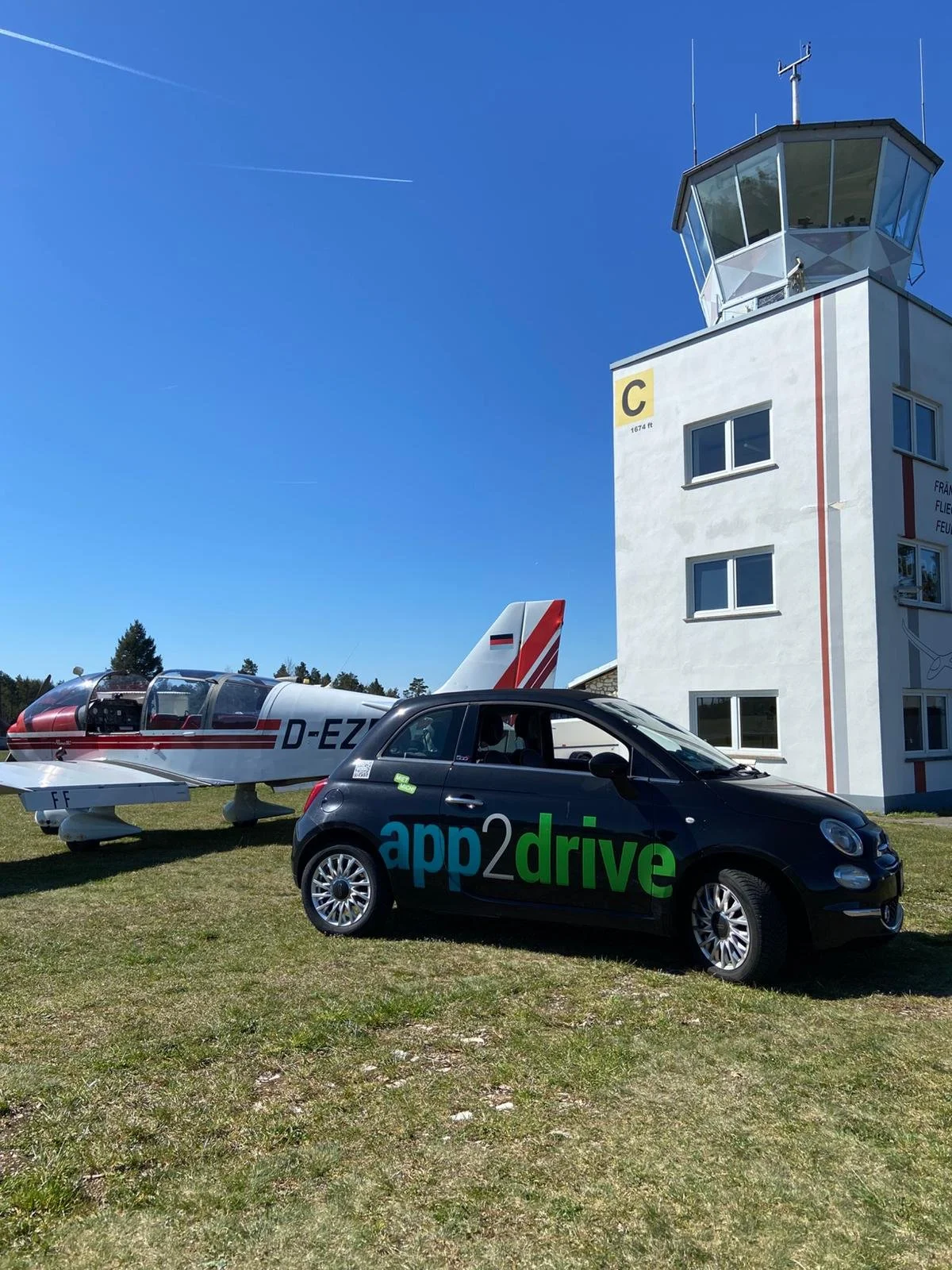Ein kleiner Jet neben einem schwarzen Auto mit dem Logo 'app2drive' vor einer Flugsicherungsturm. Das Bild ist bei sonnigem Wetter aufgenommen.
