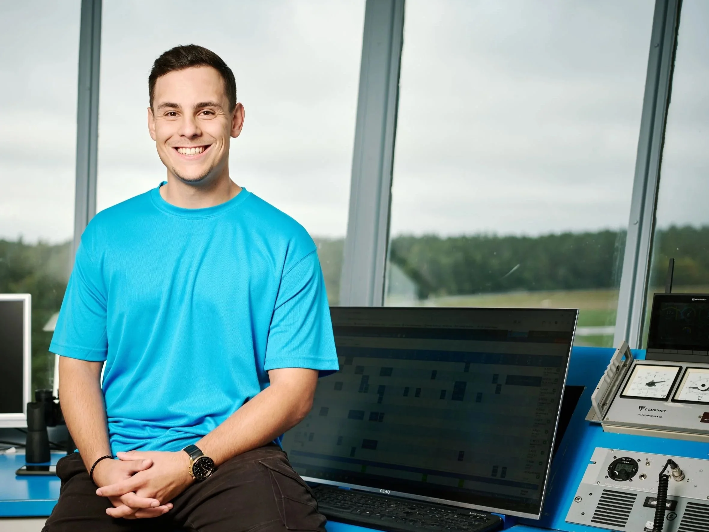 Ein junger Mann mit dunklem Haar und heller Hautfarbe, trägt ein hellblaues T-Shirt, sitzt in einem modernen Kontrollraum mit Computern und Messgeräten. Im Hintergrund große Fenster mit Blick ins Grüne bei bewölktem Himmel.