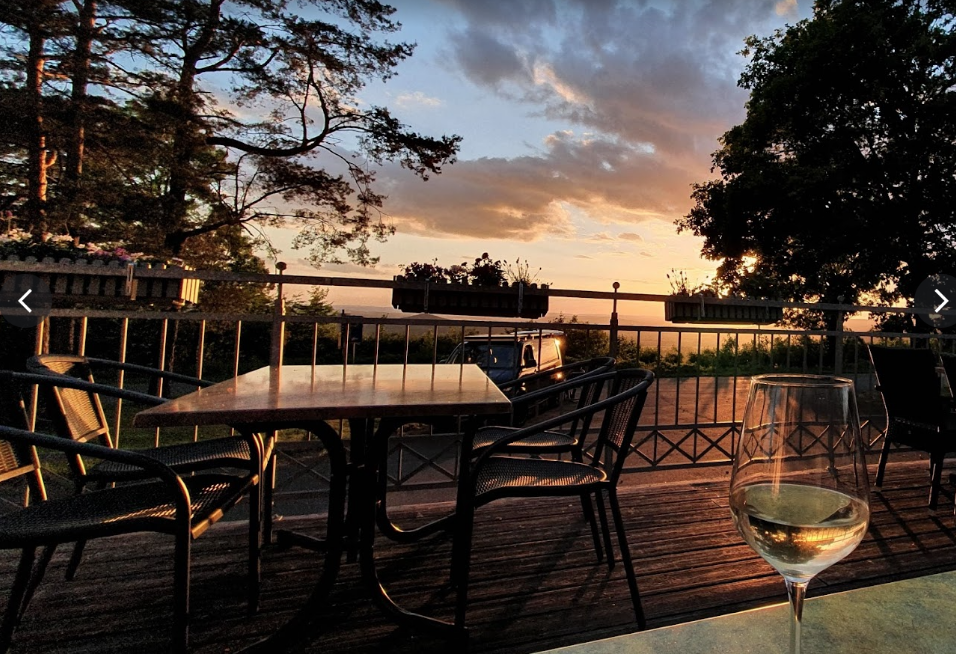Blick auf einen Sonnenuntergang von einer Terrasse mit Gartenmöbeln, einem Glas Weißwein im Vordergrund, ein Baum rechts und ein Himmel mit Wolken im Hintergrund.