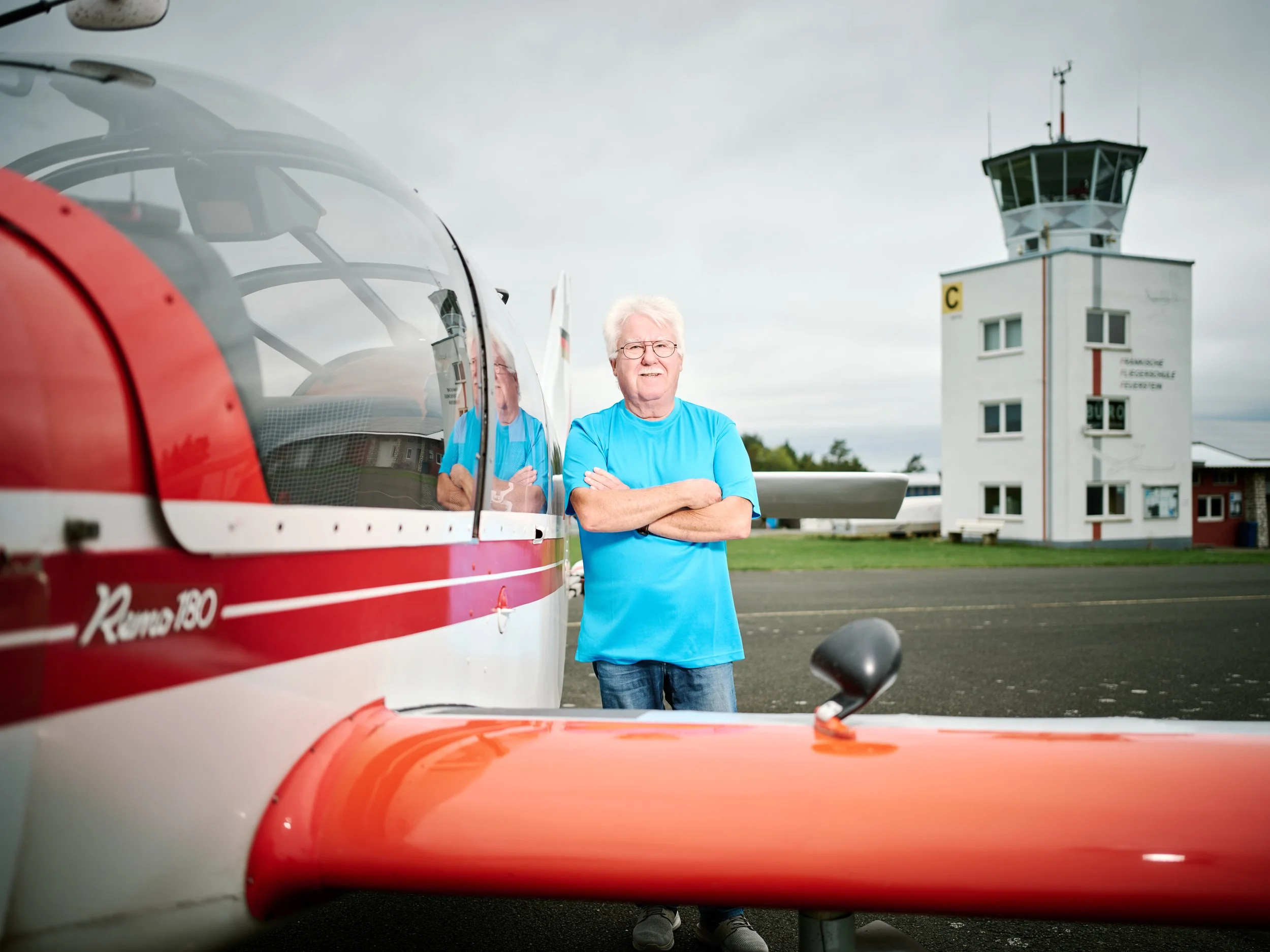 Ein älterer Mann mit weißen Haaren und Brille steht neben einem roten und weißen Flugzeug am Flughafengebäude mit einer Kontrollturm im Hintergrund.