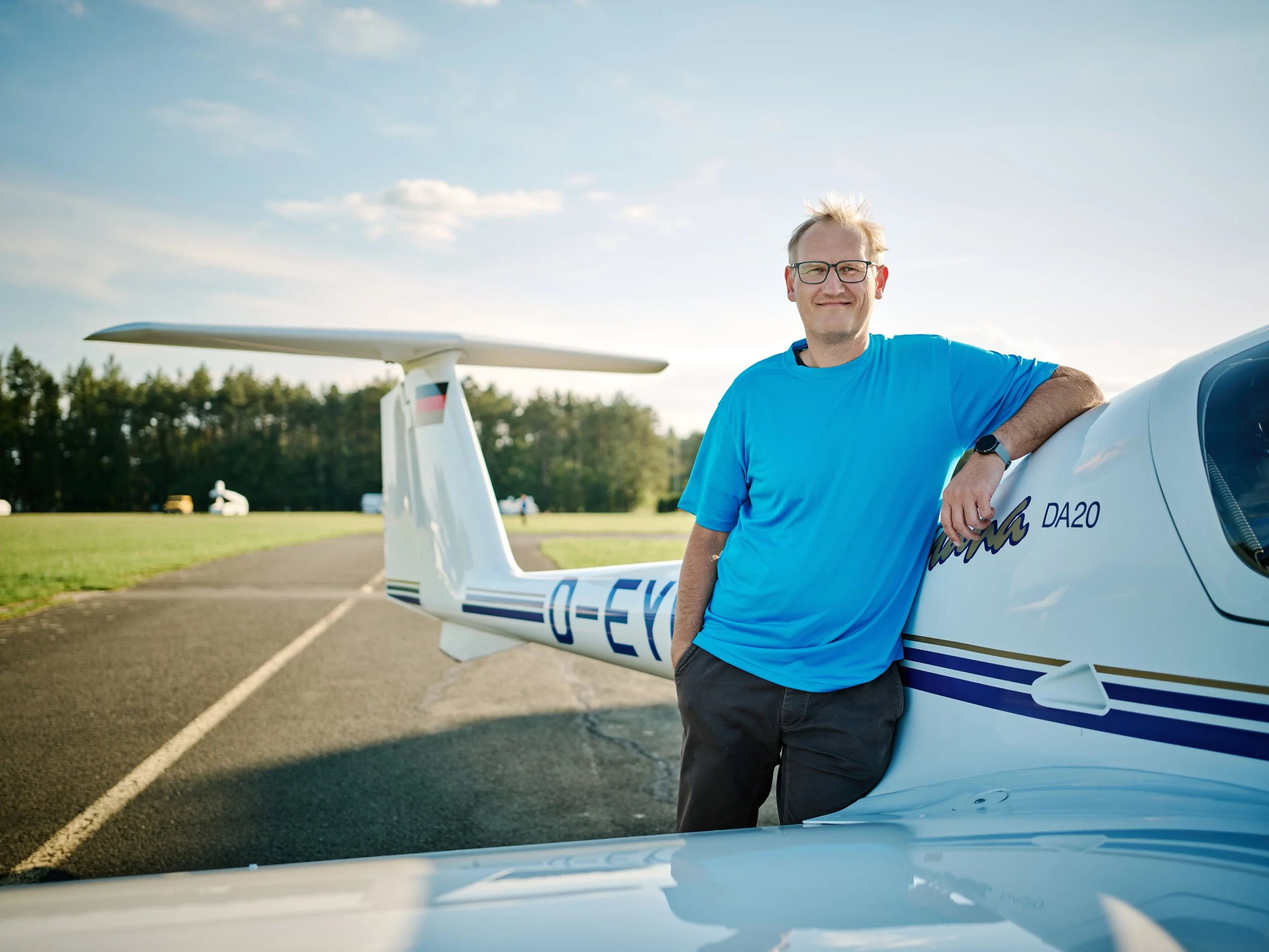 Ein Mann in einem blauen T-Shirt steht neben einem weißen Ultraleichtflugzeug auf einem Flugfeld. Im Hintergrund sind Bäume und andere Flugzeuge zu sehen, die Sonne scheint bei klarem Himmel.