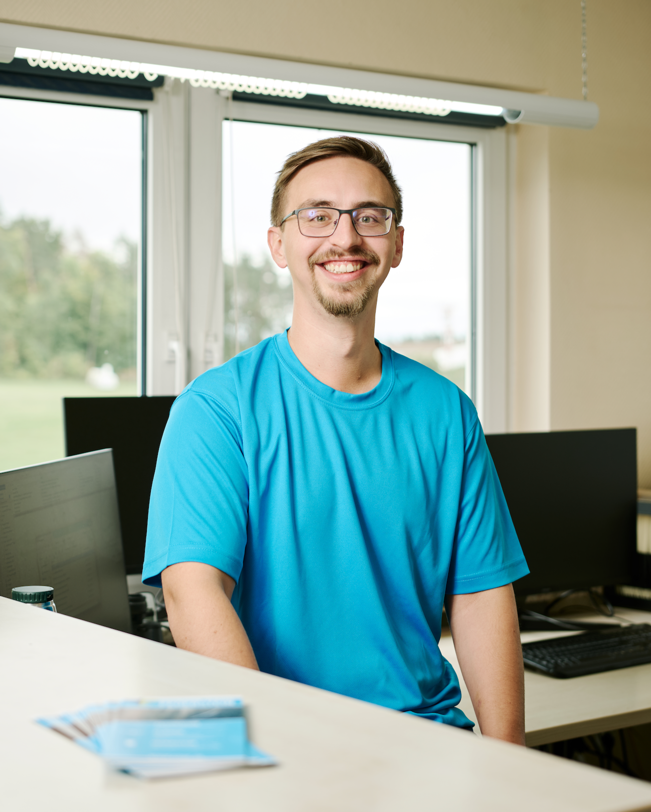 Ein junger Mann mit Brille und Bart lächelt in einem Büro, trägt ein blaues T-Shirt, hinter ihm Fenster mit Blick ins Grüne, mehrere Computermonitore auf Tischen.