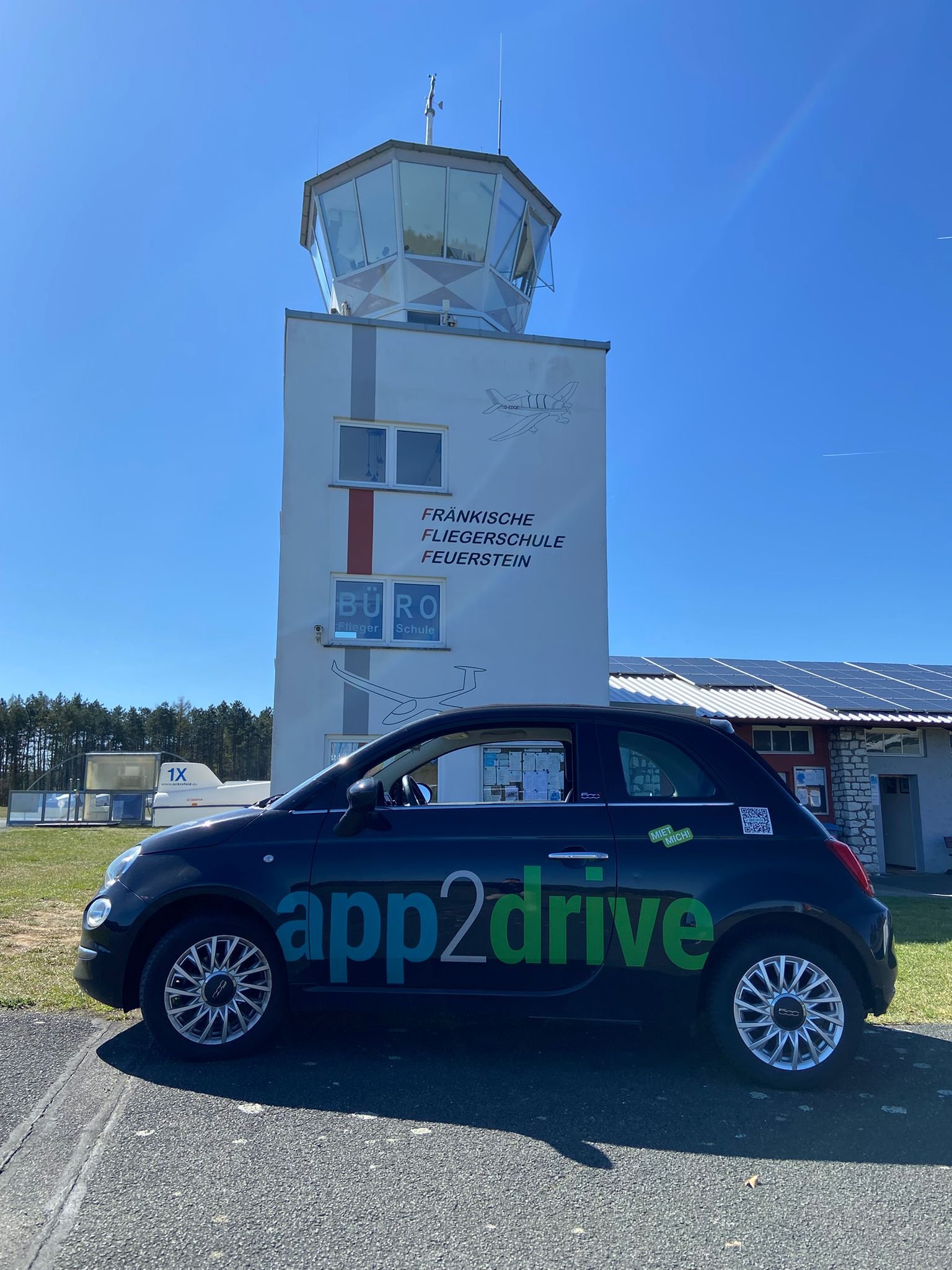 Das Bild zeigt ein schwarzes Auto mit der Aufschrift 'app2drive' in grüner und blauer Schrift. Im Hintergrund ist ein Gebäude mit einem Kontrollturm, der mit Fenstern umgeben ist, und einer Beschriftung, die auf eine Fliegerschule in Feuerstein hinweist.